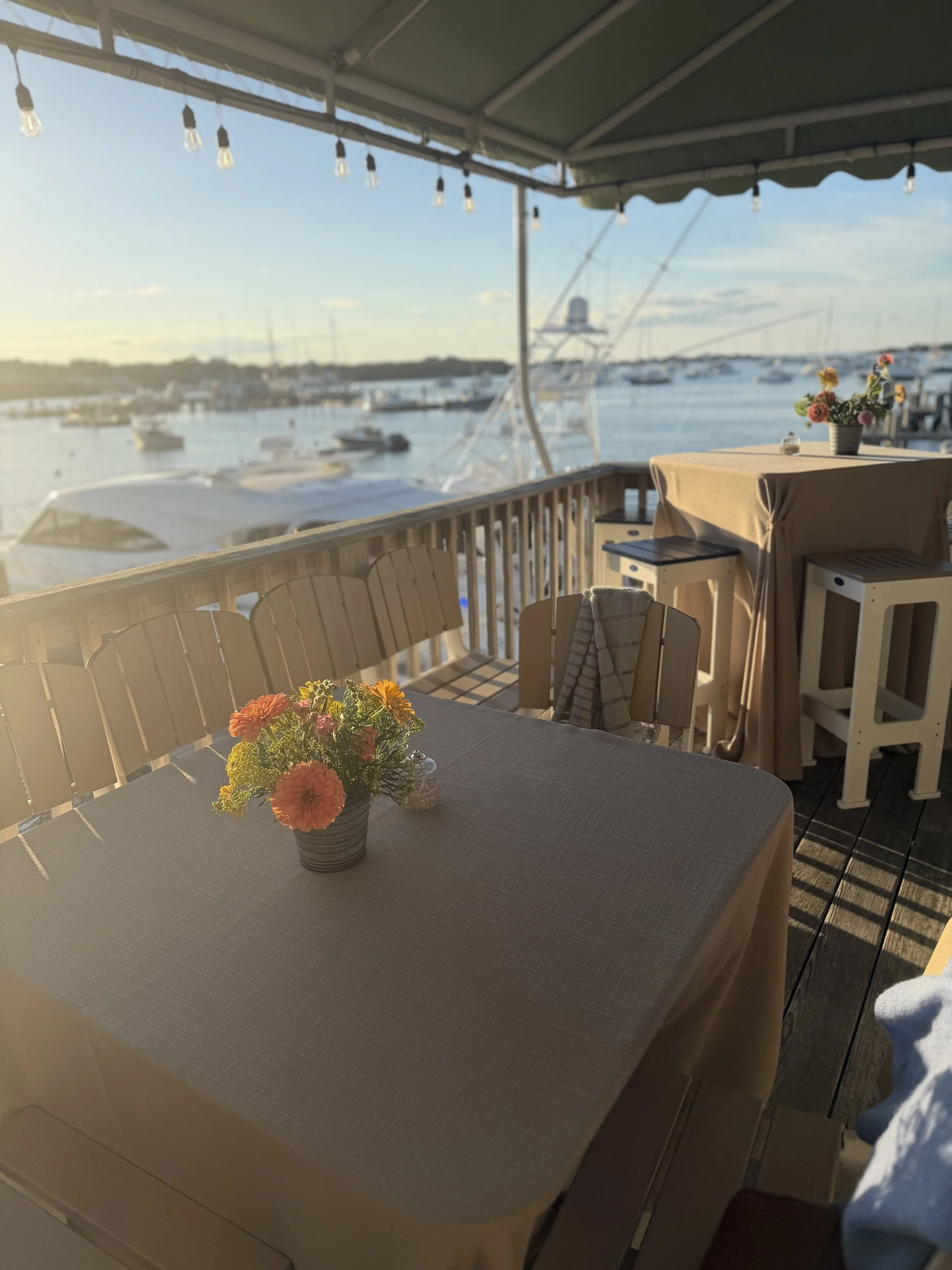 A table covered with a beige tablecloth, topped with a small flower arrangement in a gray pot, is on a balcony overlooking boats in the water during sunset, with string lights hanging above.