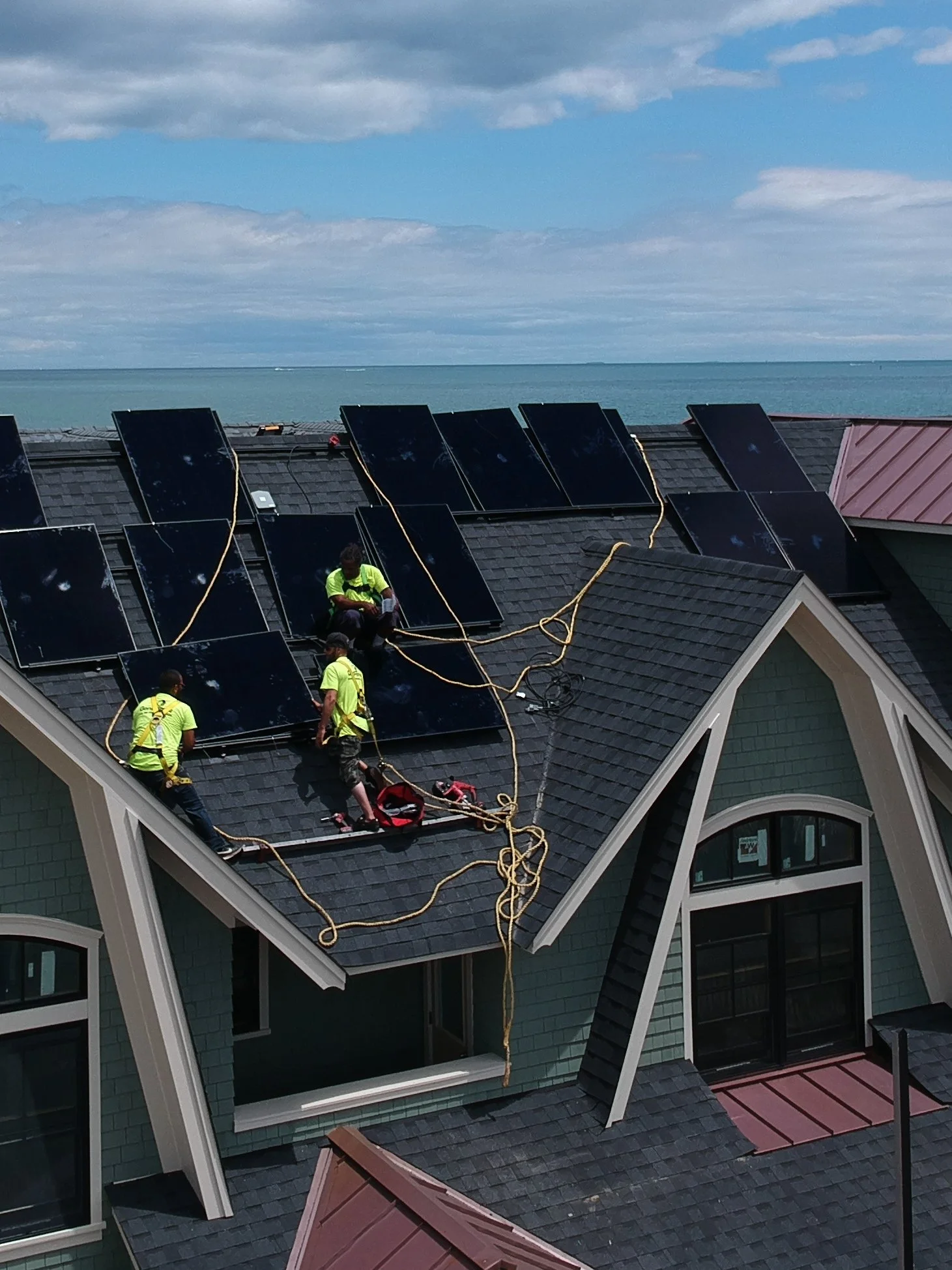 Workers installing solar panels on a rooftop of a house overlooking the ocean with a cloudy sky.