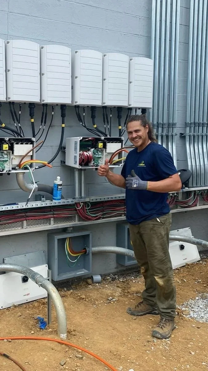 A man giving a thumbs up next to an electrical installation with circuit breakers, wiring, and other electrical components on a gray wall.