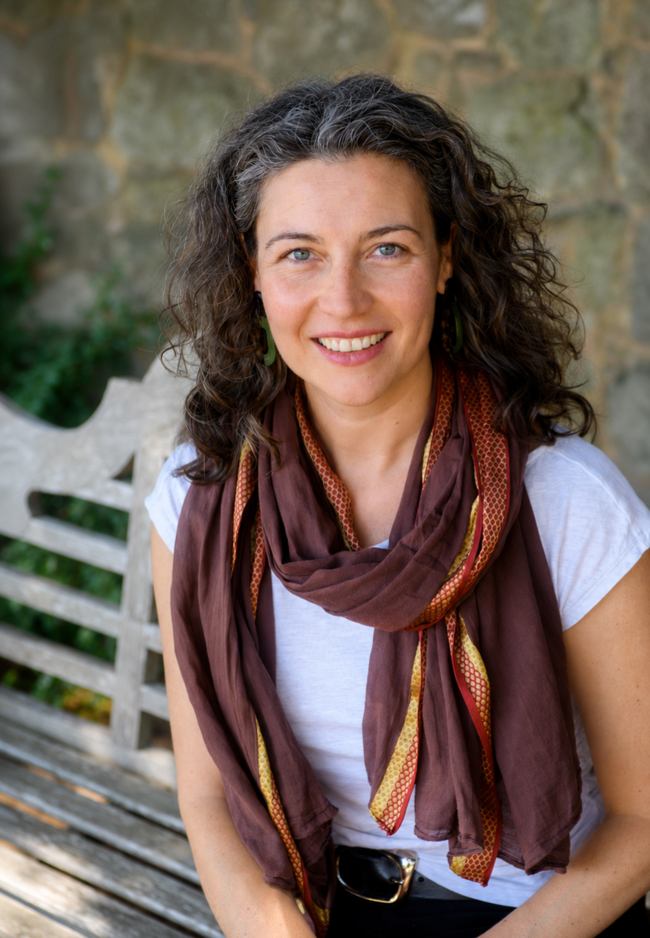 Portrait of Anne-Julie Burdet, independent consultant. A woman with curly brown hair, blue eyes, and wearing a white t-shirt and a brown scarf with gold accents, sitting on a wooden bench outdoors with a stone wall and greenery in the background.