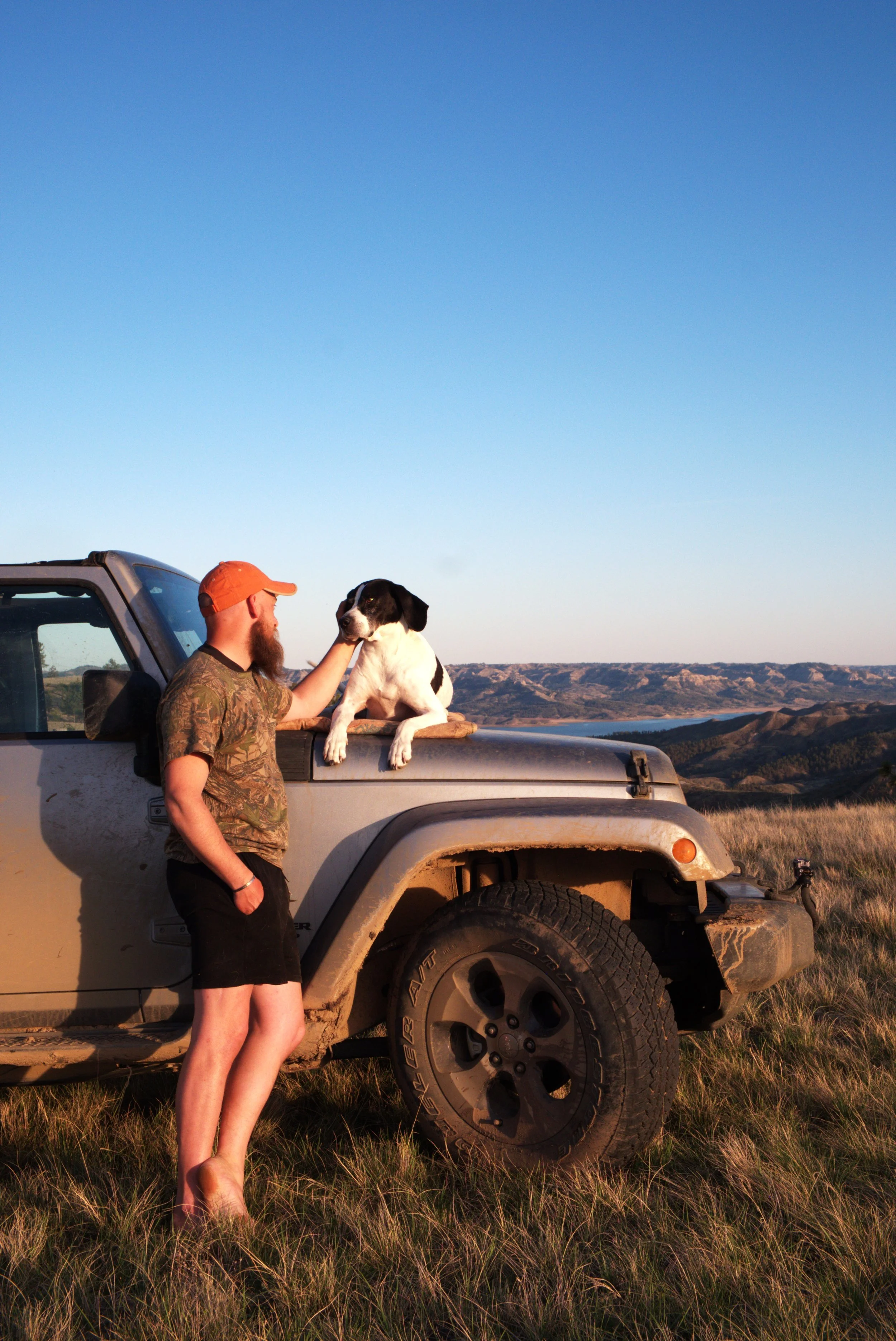 A man with a beard, wearing an orange cap, a camouflage t-shirt, and black shorts, standing next to a beige off-road vehicle in a grassy field during sunset. The man is petting a large black and white dog that is sitting on the vehicle's hood, with a landscape of hills and water in the background under a blue sky.