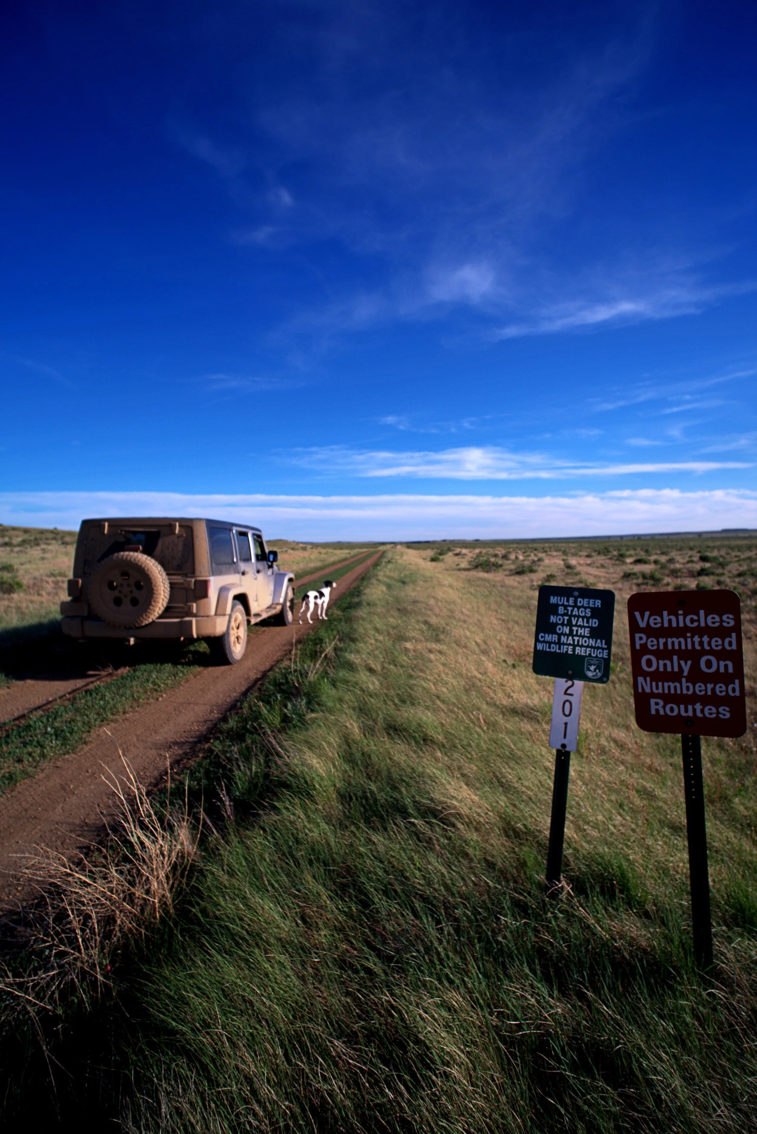 A dusty off-road vehicle parked on a grassy trail in a wide open landscape under a deep blue sky with scattered clouds. There is a sign in the foreground that reads 'Vicle Permitted Only On Numbered Routes' and another sign about mule deer tags and wildlife refuge.