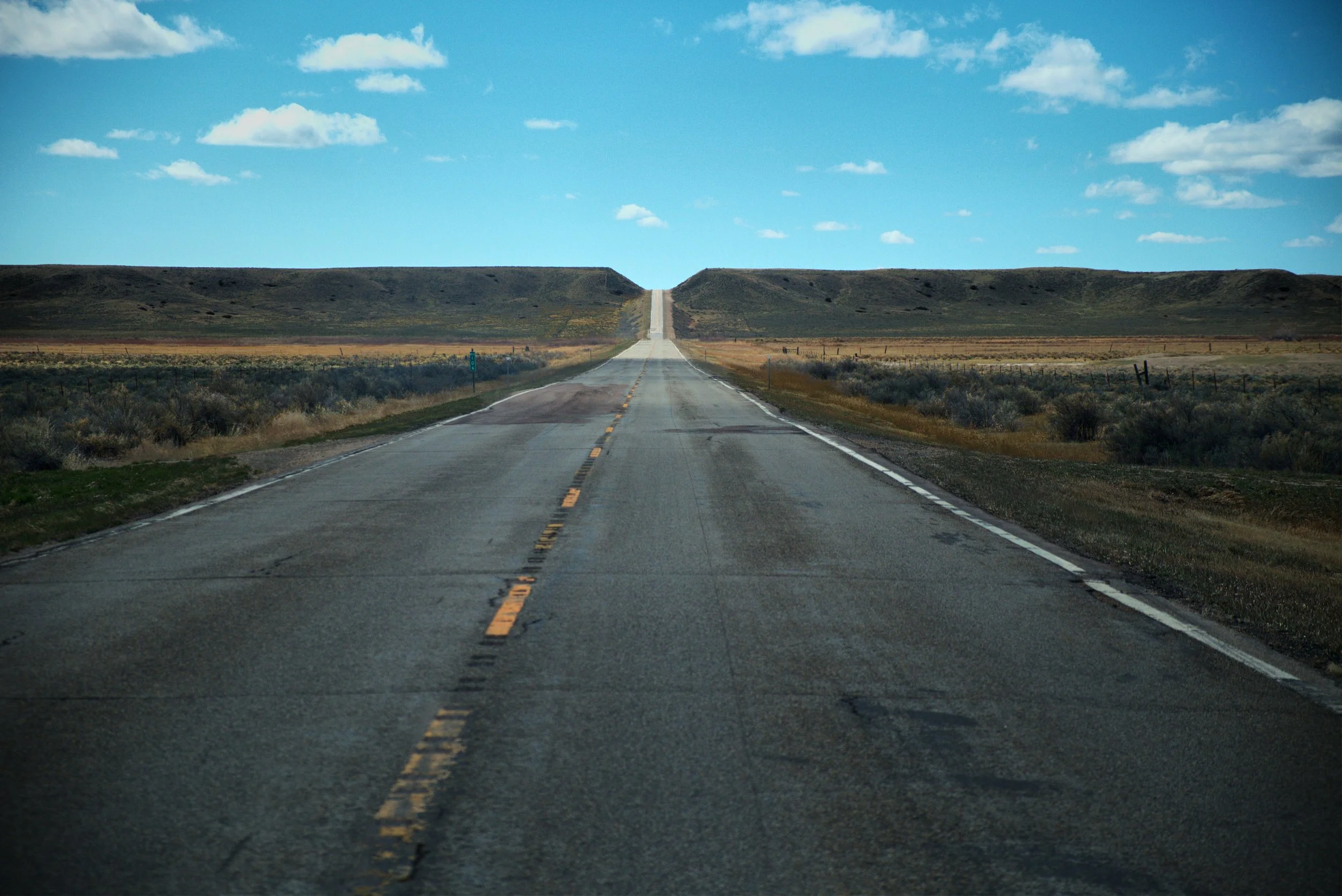 Long empty road stretching into hills under blue sky with clouds.