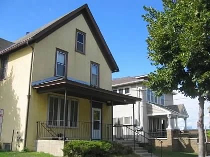A yellow two-story house with a porch, railing, and stairs, surrounded by a lawn and trees, under a clear blue sky.