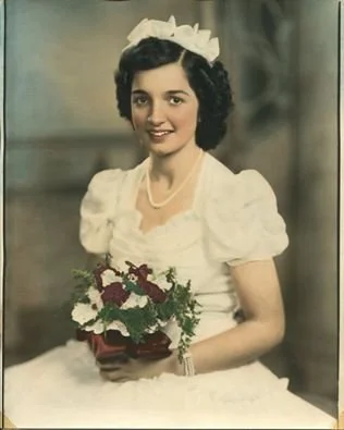 A woman with short dark curly hair dressed in a white dress, wearing a white bow headband, holding a bouquet of white and purple flowers, and smiling at the camera.