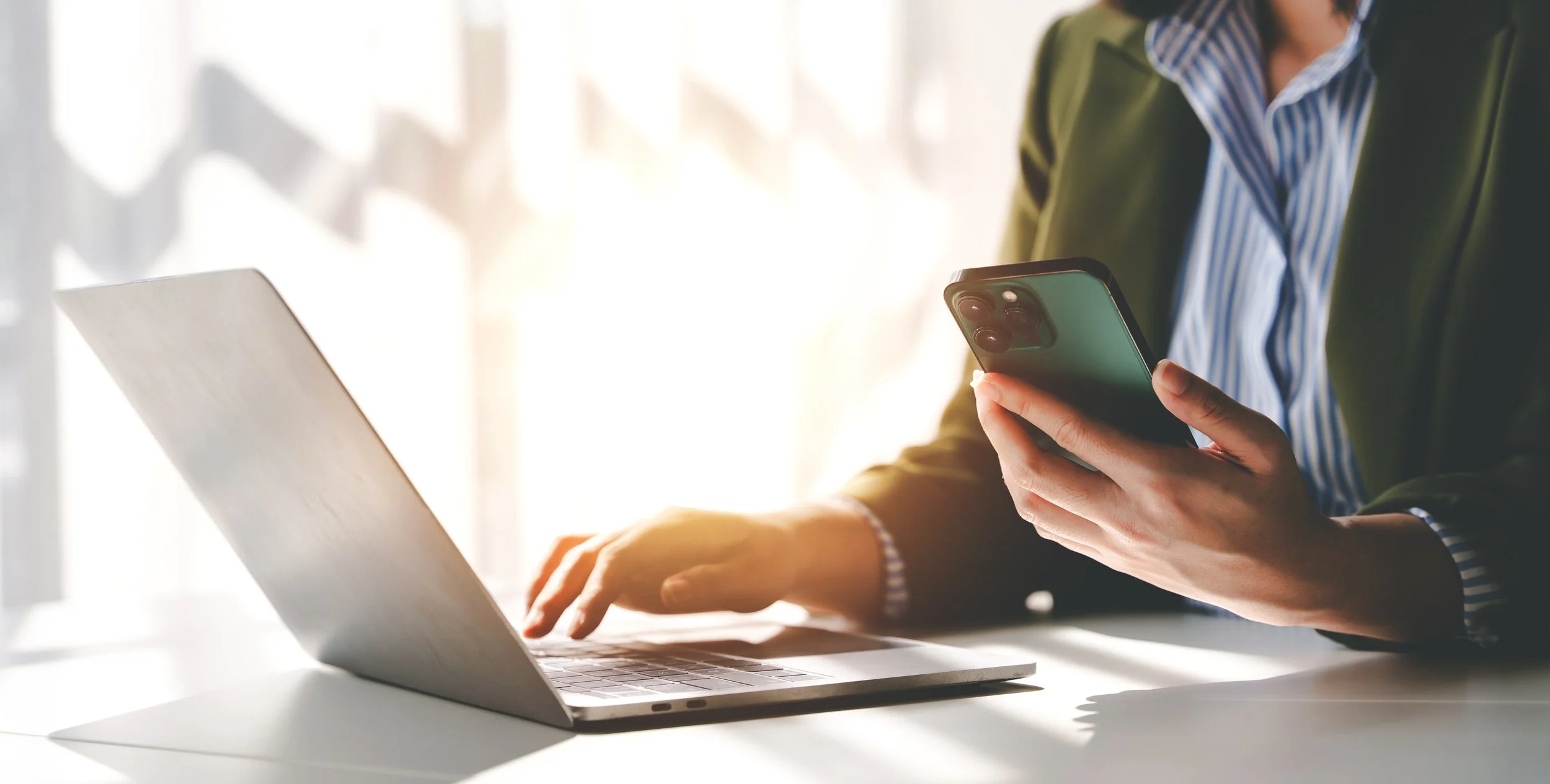 Person working on a laptop and holding a smartphone on a desk with sunlight shining in the background.
