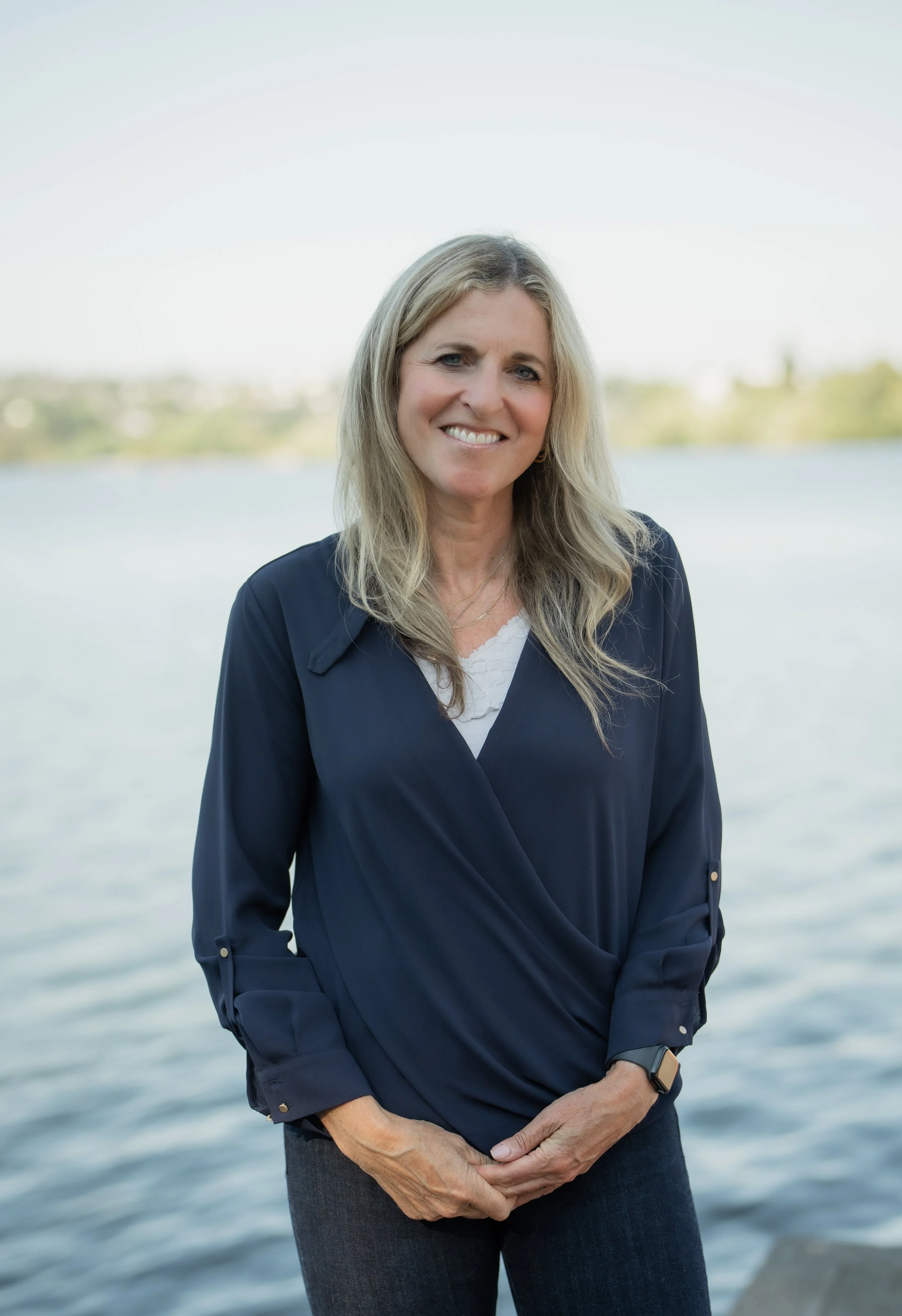 A woman with long blonde hair smiling outdoors by water, wearing a navy blue jacket and dark jeans.