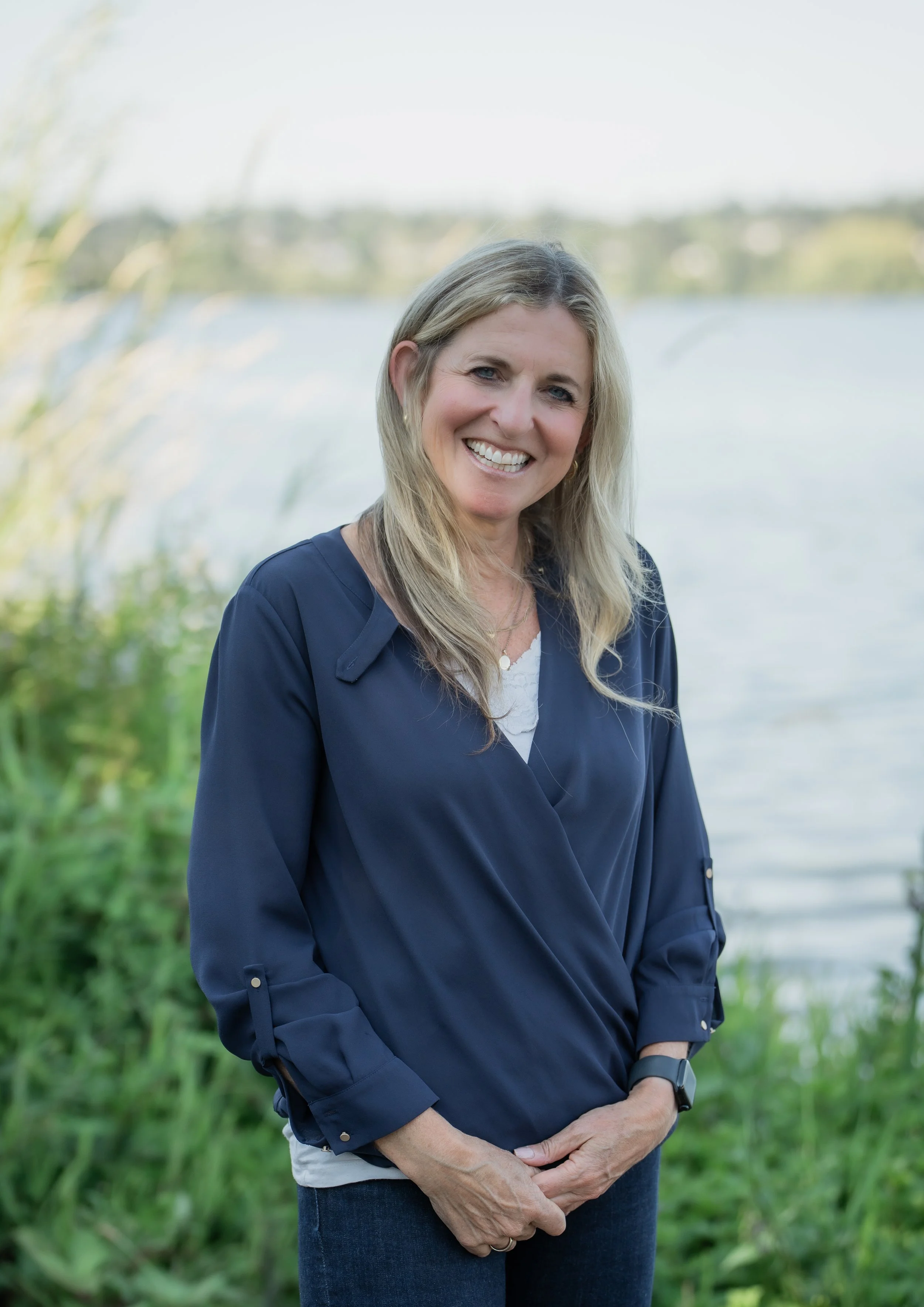 A smiling woman with blonde hair, blue eyes, and light skin, standing outdoors near a body of water with greenery and trees in the background, wearing a navy blue shirt and a watch on her left wrist.