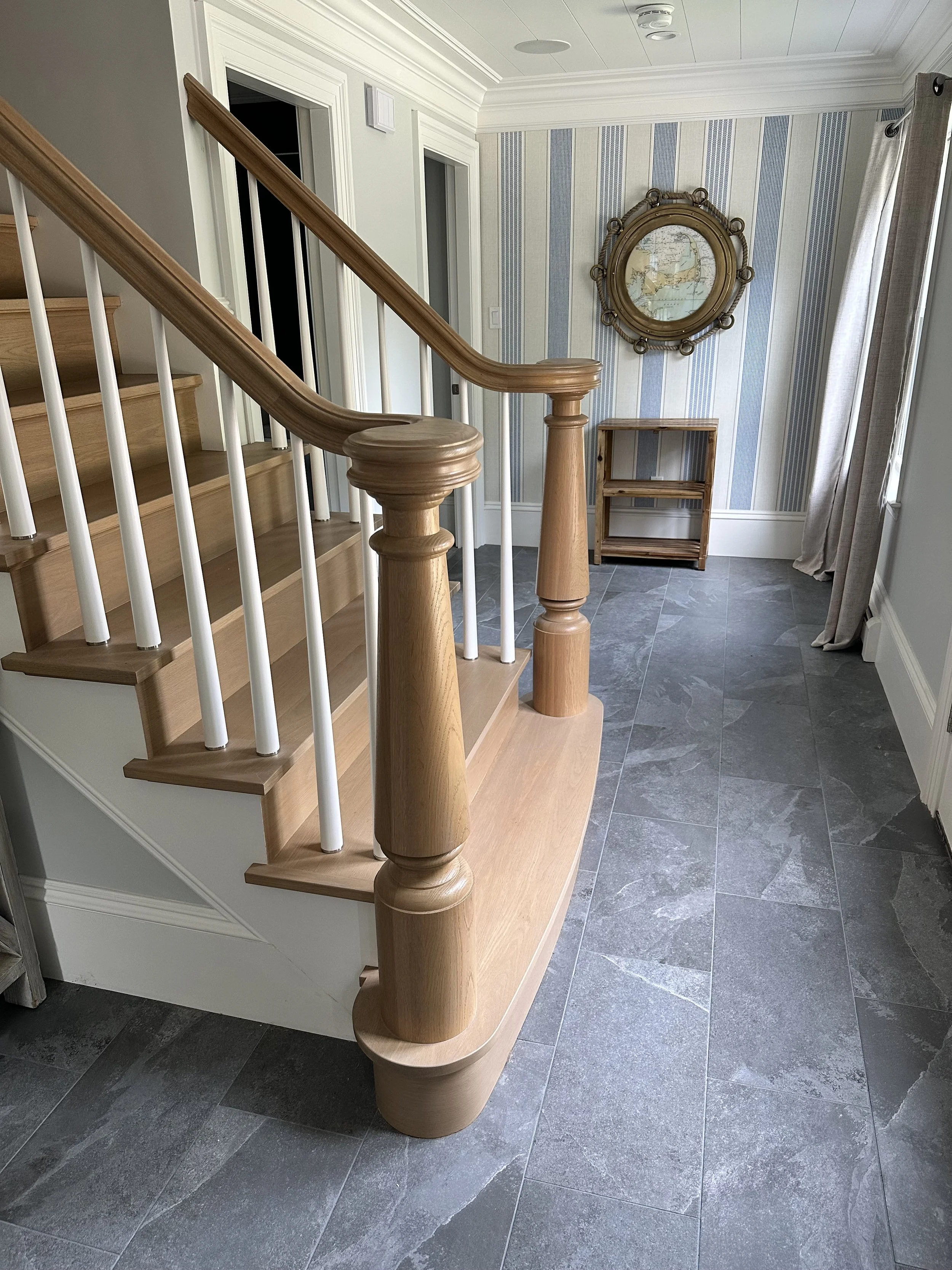 A staircase with a rounded wooden handrail and white spindles, leading to an upper floor. The wall behind has blue and beige striped wallpaper, with a large circular mirror with a rope design frame, and a small wooden shelf underneath. To the right, 