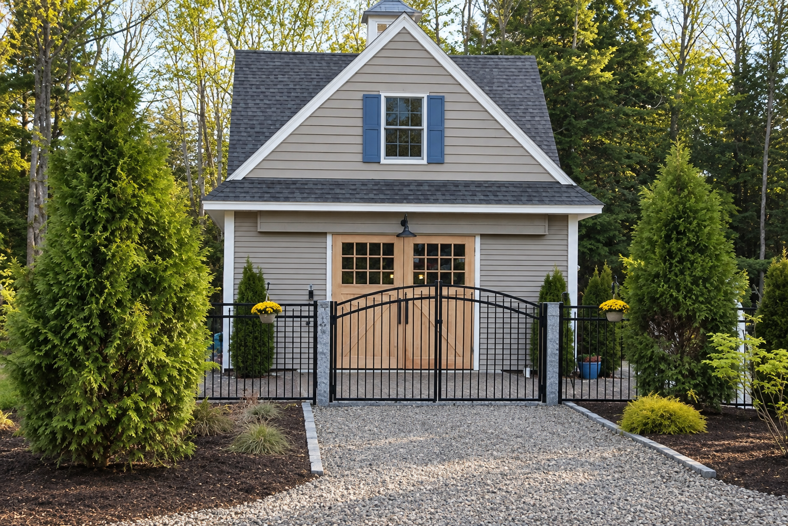A quaint house with beige siding, a gray shingled roof, blue shuttered window, and wooden double doors, surrounded by black metal fencing with yellow flowers on the gate and lush green trees in the background.