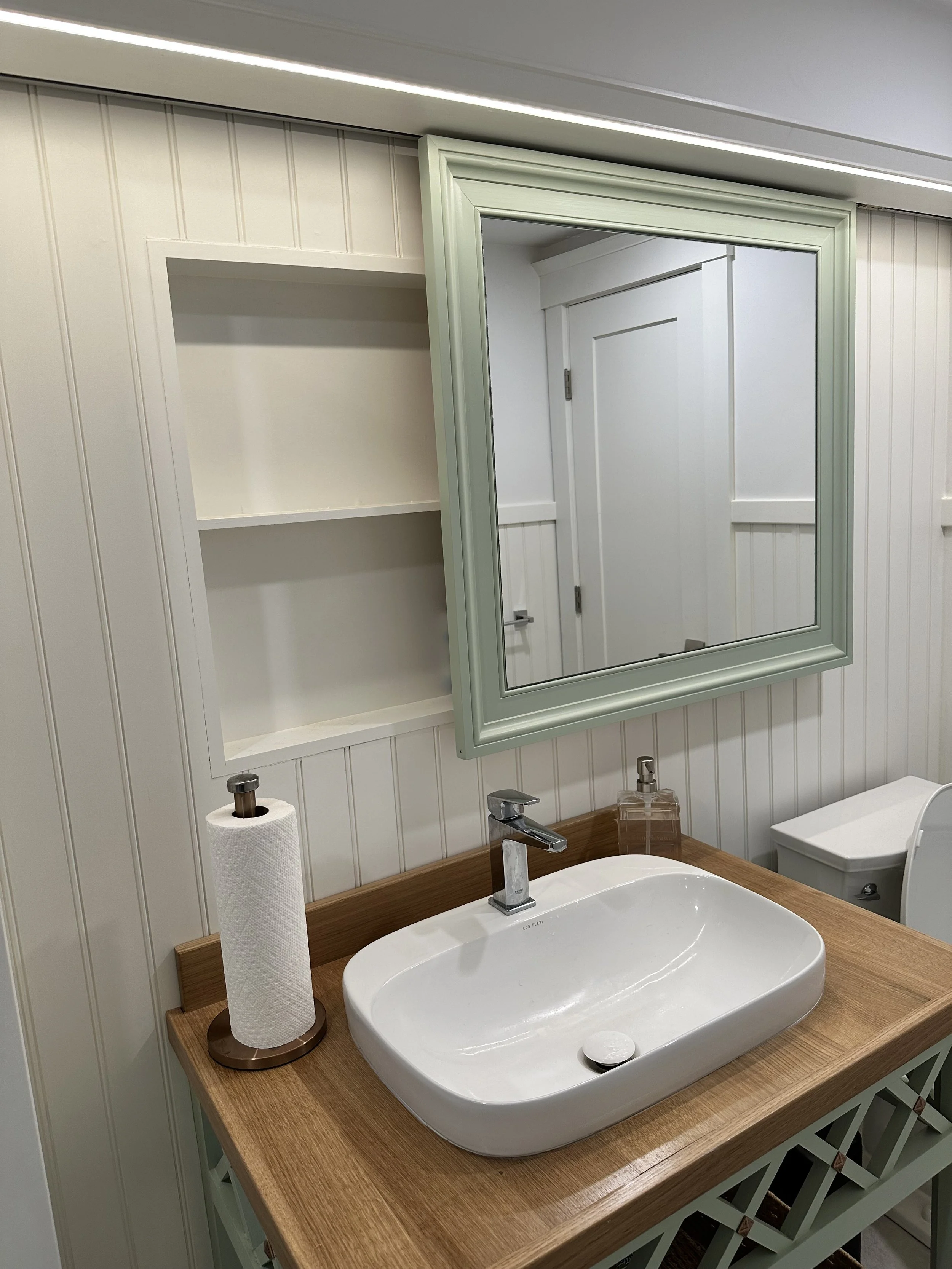 A bathroom vanity area with a white sink, a silver faucet, a paper towel roll on a holder, a soap dispenser, a mirror, and a white toilet in the background. The counter has a wooden surface and the wall has white beadboard paneling.