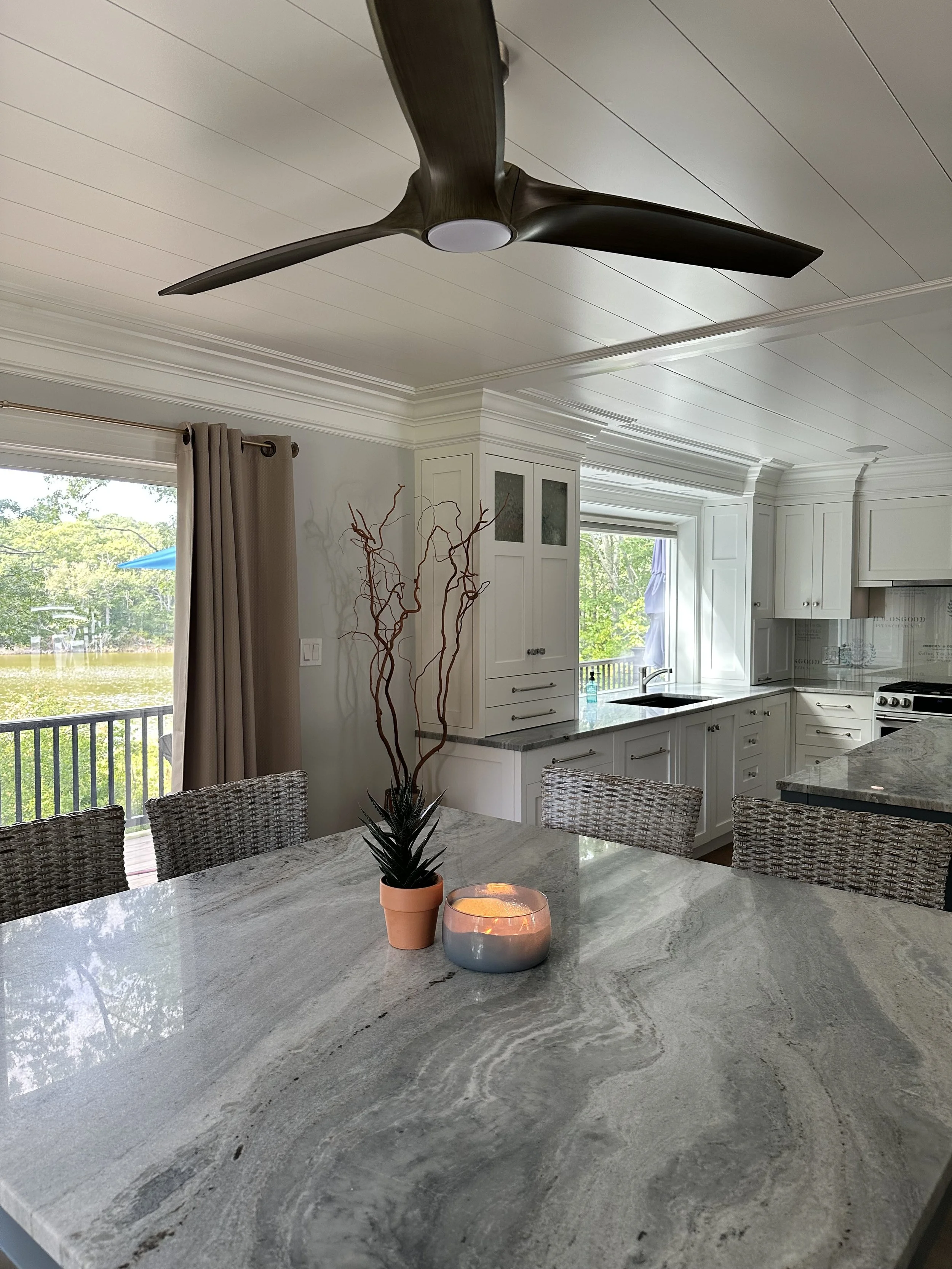 A kitchen with white cabinets, a large window, and a gray marble countertop. There is a potted plant and a pink candle holder with a lit candle on the counter. A ceiling fan hangs from the ceiling, and a dining area with wicker chairs is visible.