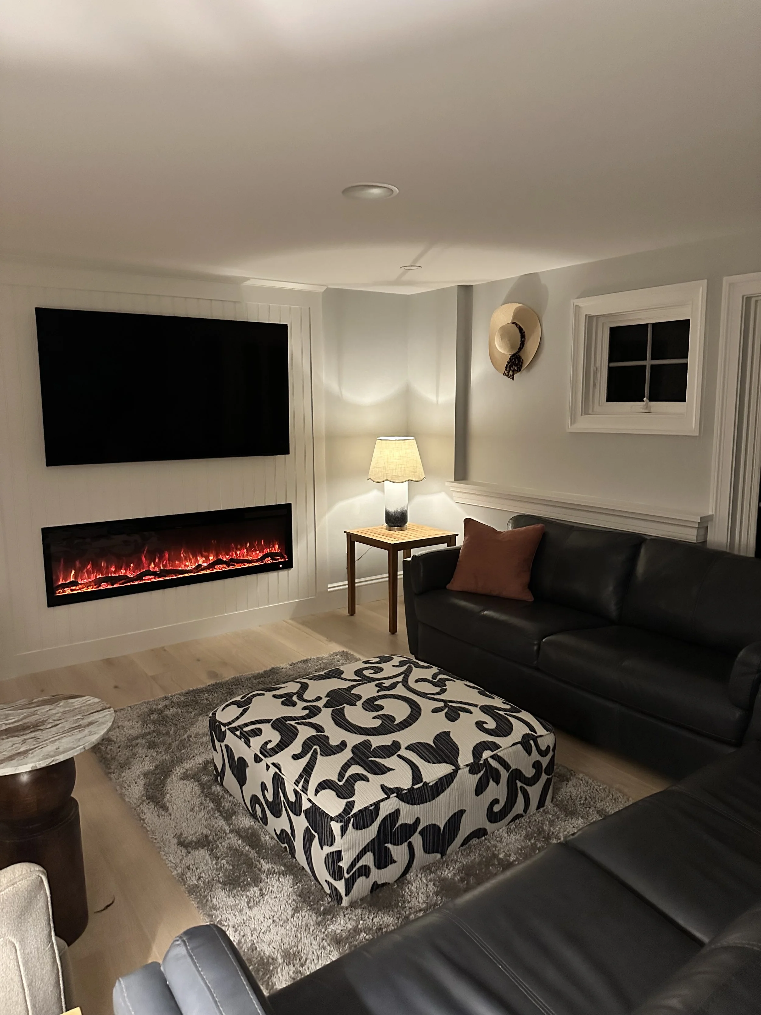 Living room with a black leather sofa, an upholstered ottoman with a black and white abstract pattern, a small round marble side table, a lamp with a beige shade, a wall-mounted TV, a wall-mounted electric fireplace, a beige hat with a scarf hanging 