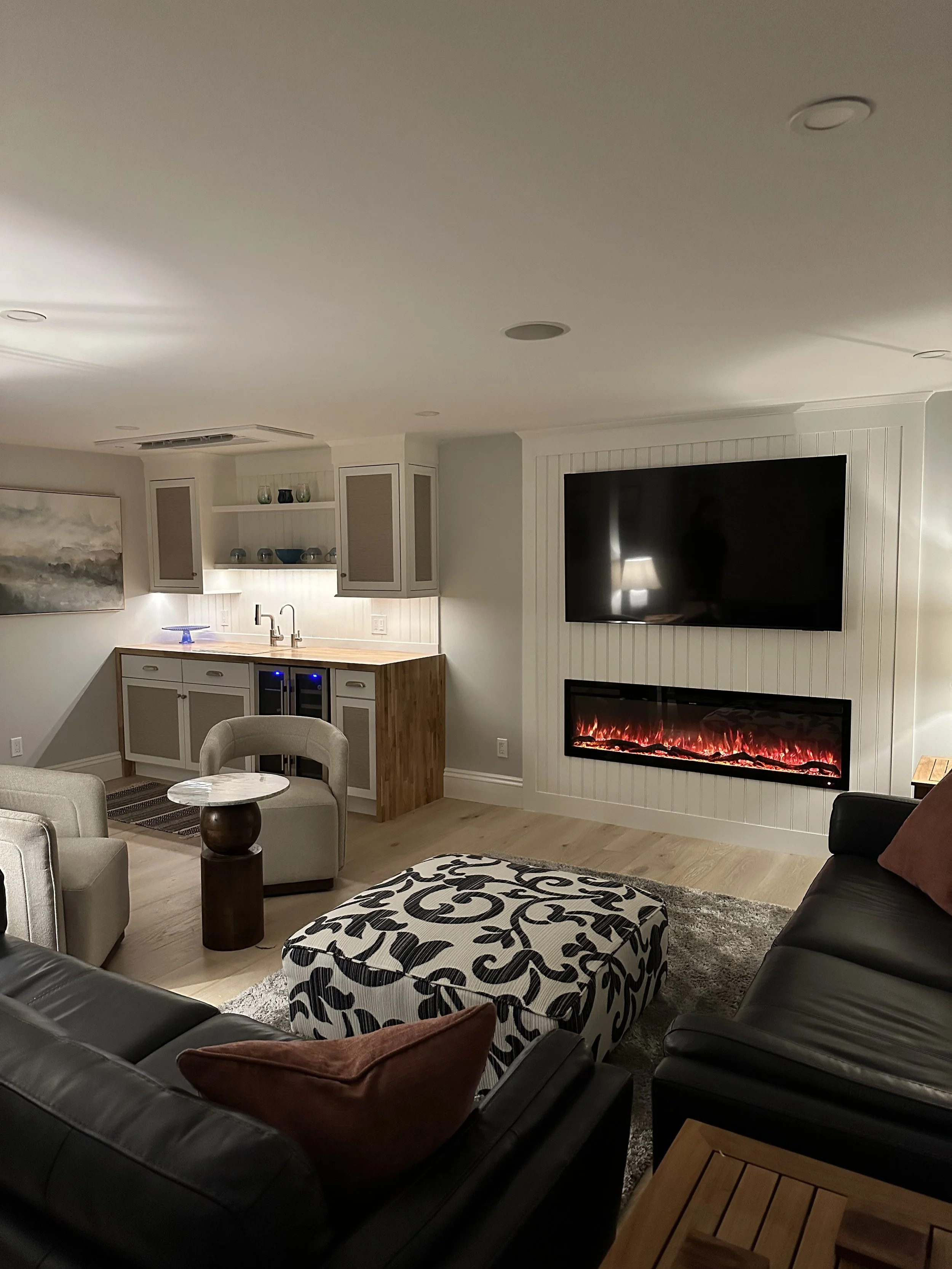 Living room with black leather sofa, beige armchair, ottoman with black and white pattern, small round table, fireplace, television, and kitchen area with white cabinets and wooden countertop.