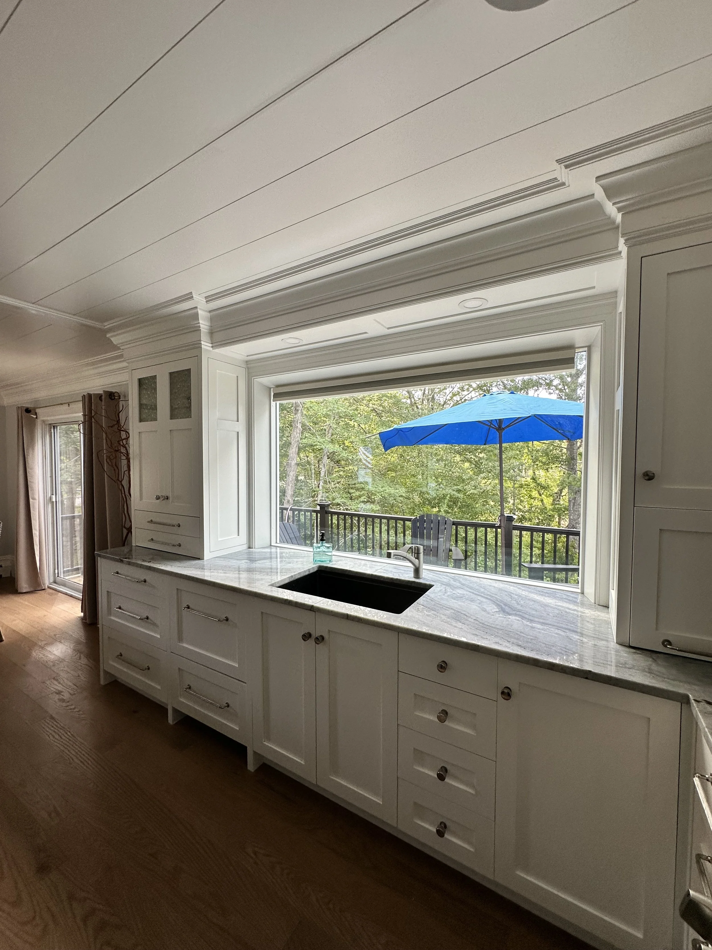 White kitchen cabinet with marble countertop and a large window showing trees outside. There is a blue umbrella outside on the patio.