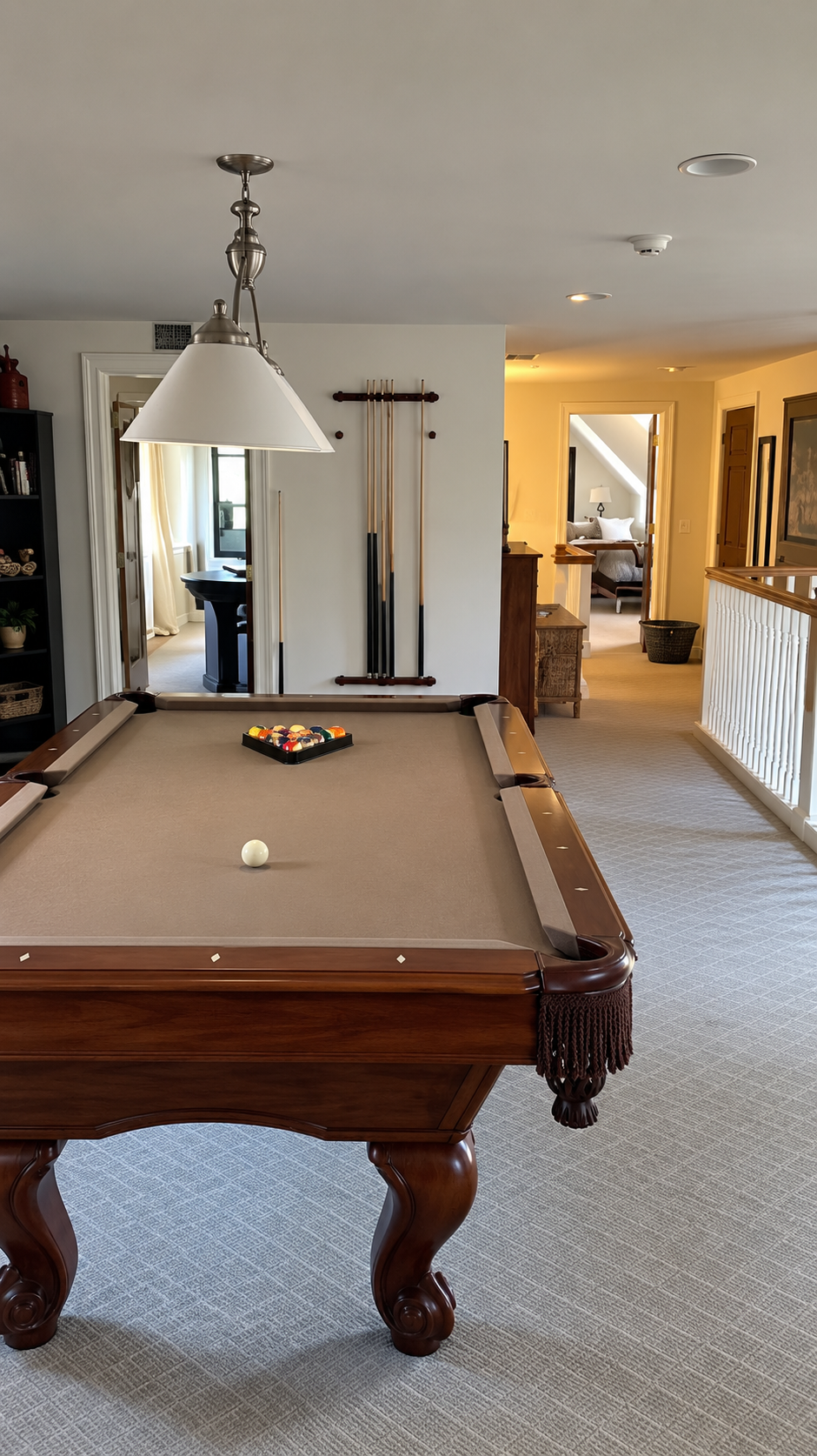 Photo of a pool table in a home game room with a white ball and a tray of pool balls, pool cues on the wall, and an open hallway leading to bedrooms.