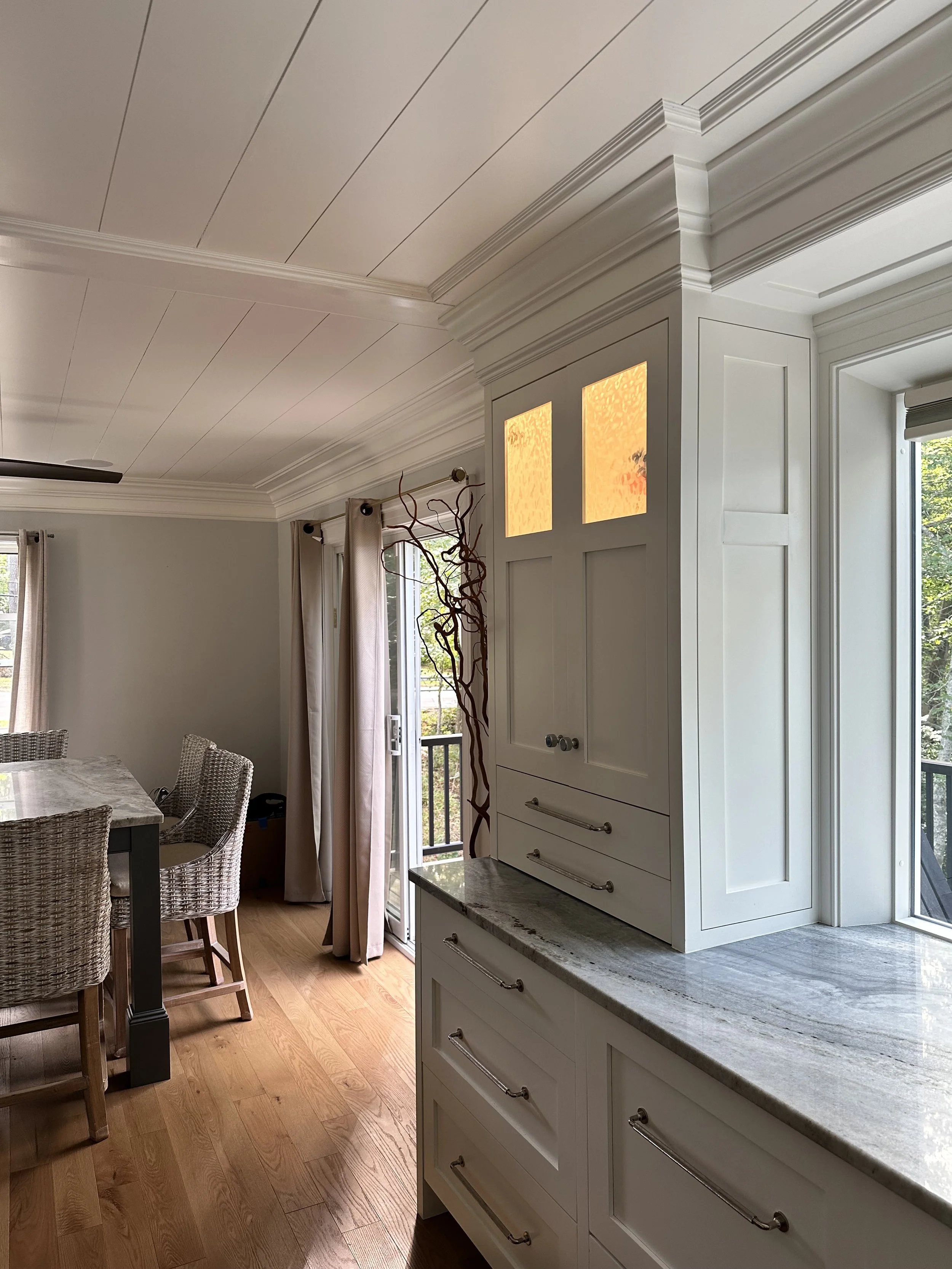 A bright kitchen corner with white cabinetry and granite countertops, hardwood floors, a dining table with wicker chairs, a sliding glass door with curtains, and a decorative branch in a vase.