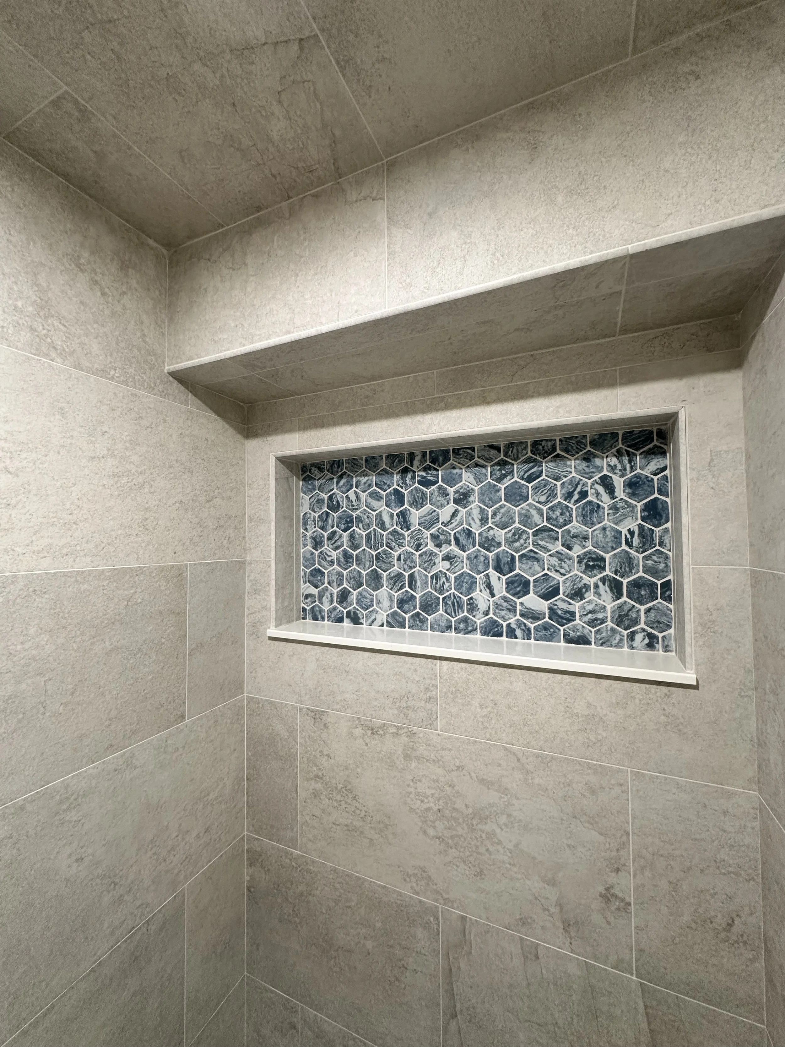 Close-up of a shower wall with gray stone tiles, a niche with hexagonal blue and white marble tiles, and a corner with a shelf or ledge.