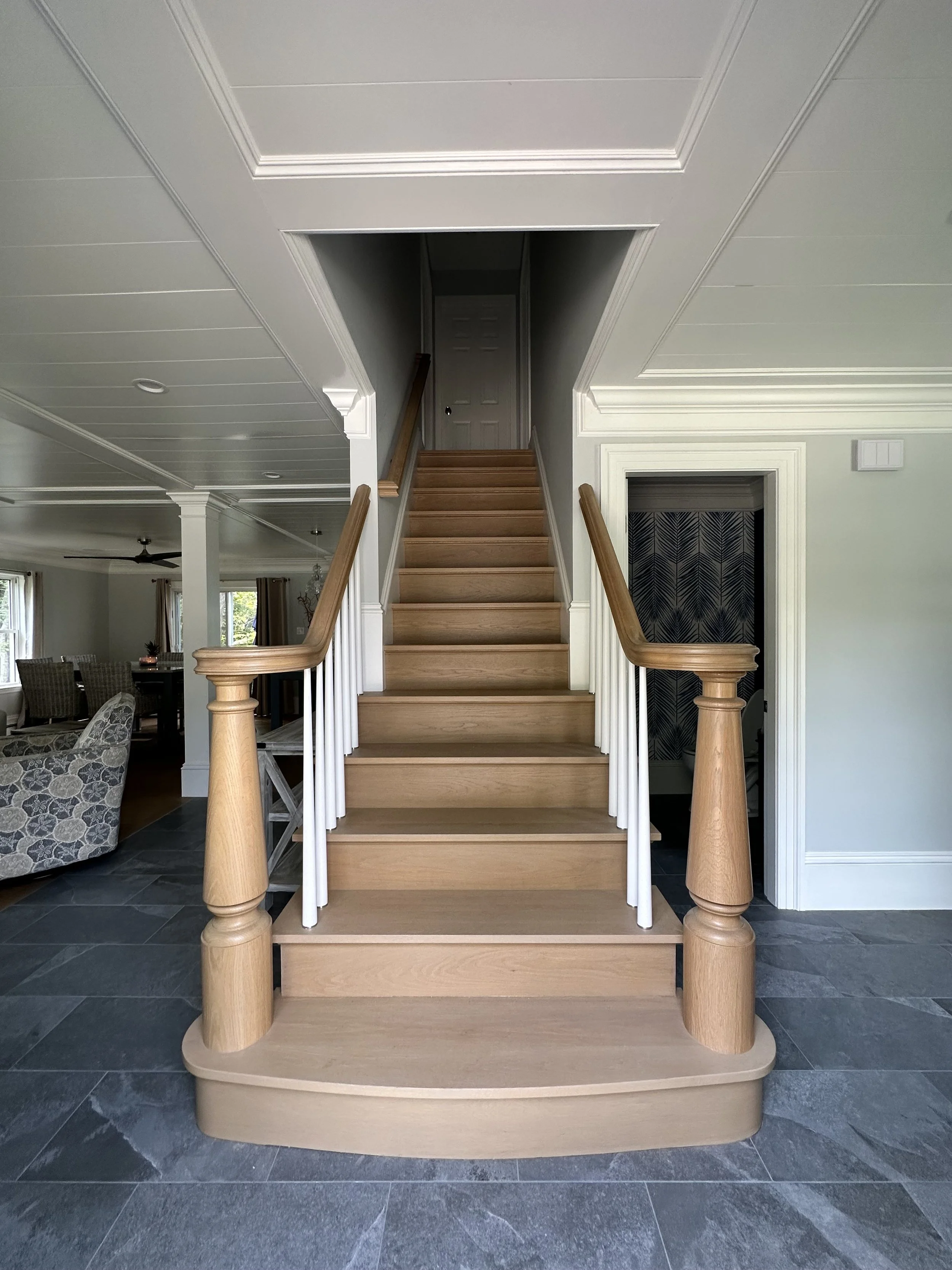 Wood staircase with a rounded base and wooden handrails, leading to a closed door at the top, in a room with light-colored walls and dark tiled flooring.