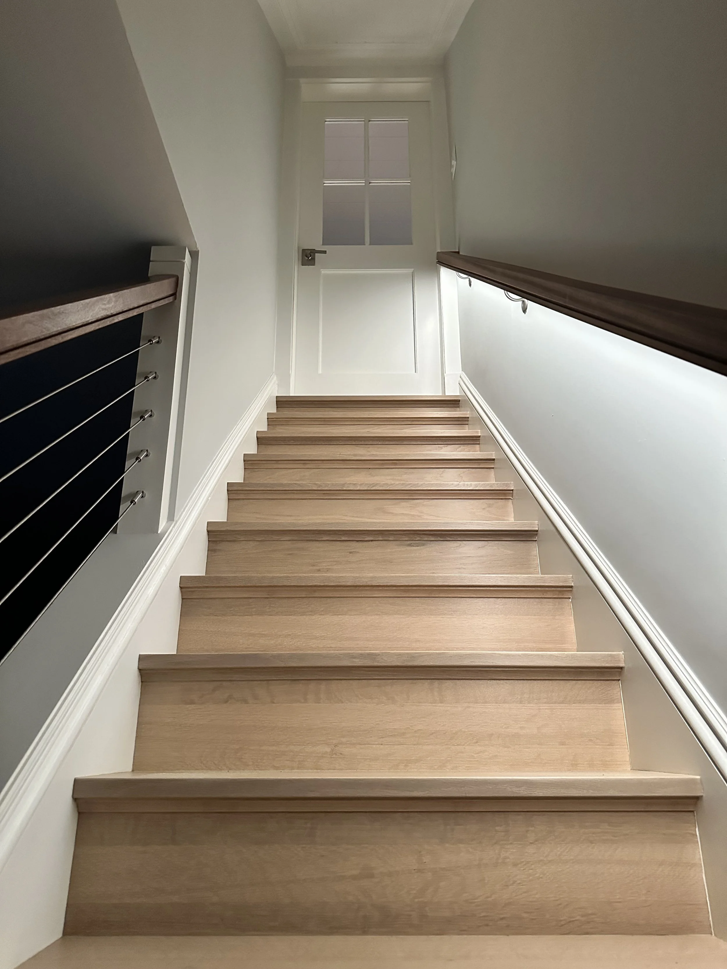 Indoor staircase with wooden steps, white walls, and a white door at the top, viewed from the bottom of the stairs.