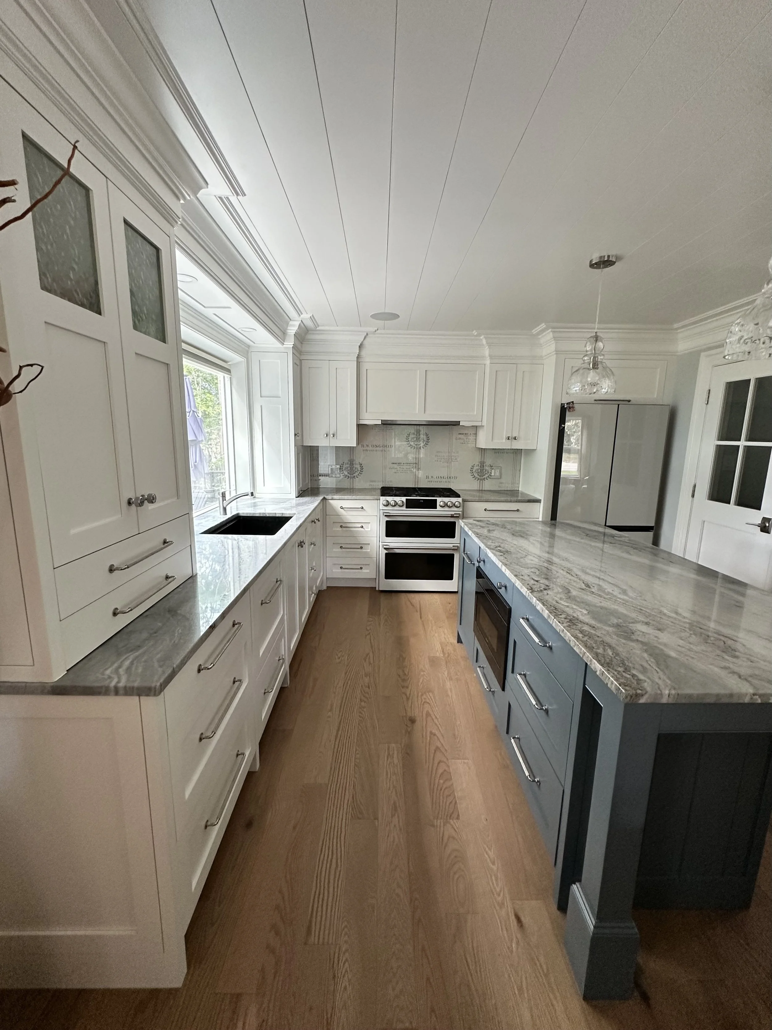 A kitchen with white cabinetry, a large window, a marble countertop island, a stainless steel oven, and hardwood floors.
