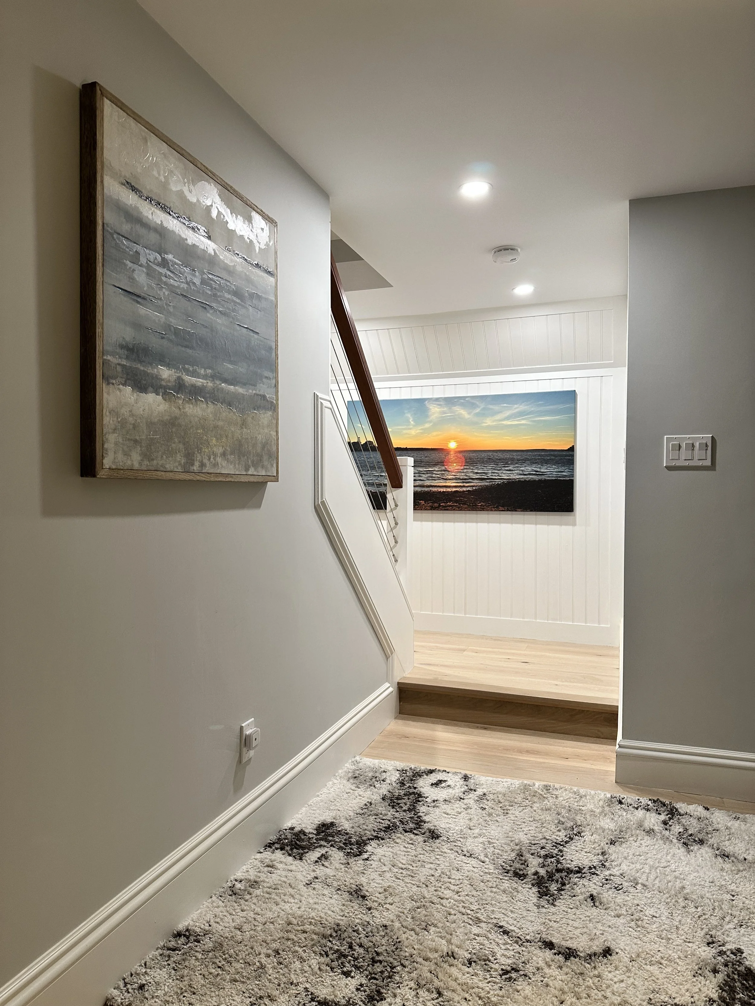 An indoor hallway with a large abstract painting on the left wall, a short staircase leading to a room with a wall-mounted art of a sunset over the ocean, wood flooring, a shaggy rug, and ceiling lights.