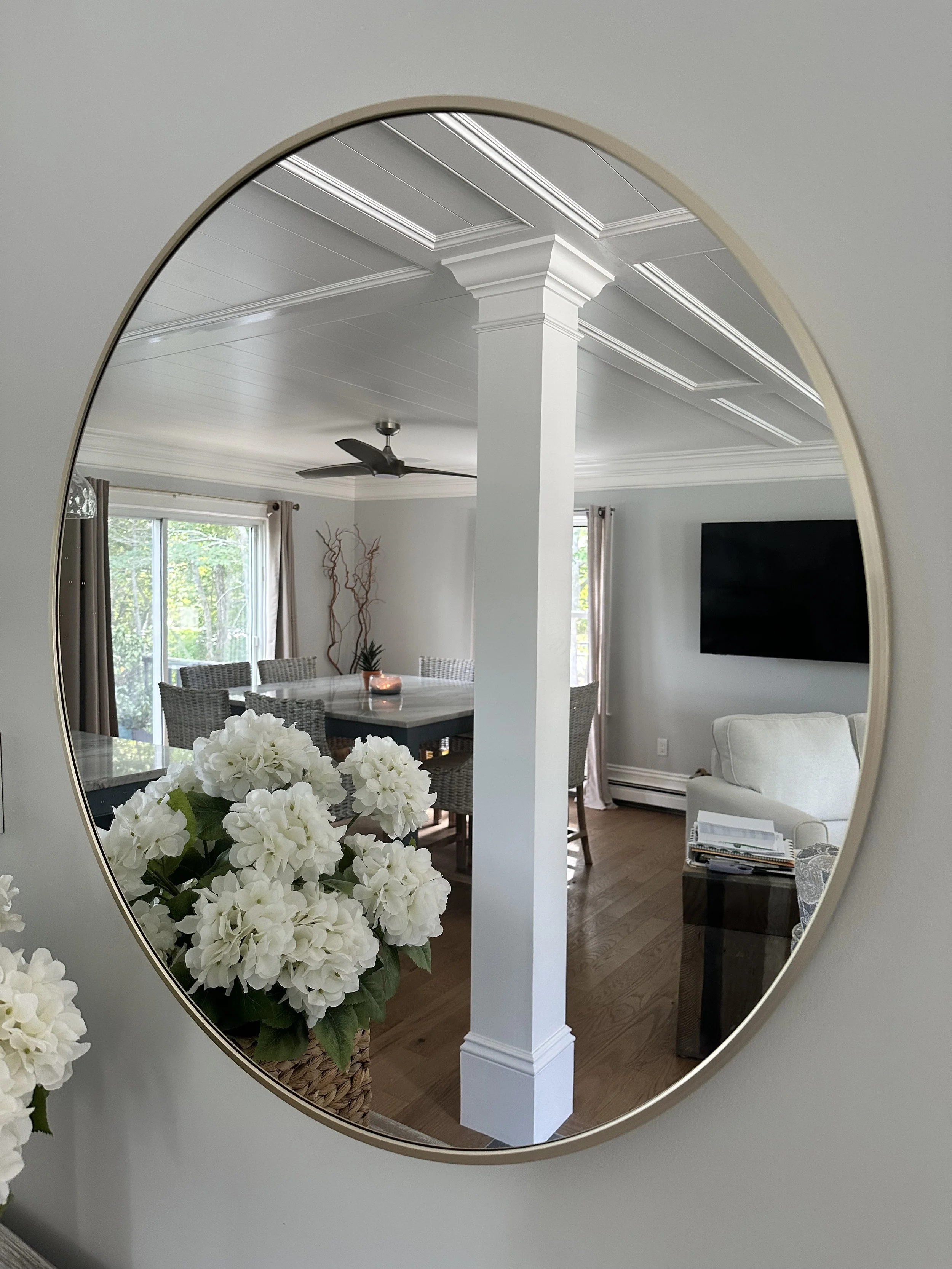 Interior view of a living and dining area seen through a circular mirror. The room has large windows, a ceiling fan, a white column with molding, and a flat-screen TV mounted on the wall. There are white flowers in a basket in the foreground.