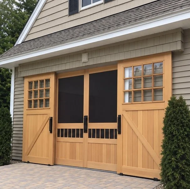A house with a large wooden garage door that has a sliding barn door style. The garage door features two small windows on each side and a black sliding gate in the middle. The house has beige siding, and there are two tall green bushes flanking the g