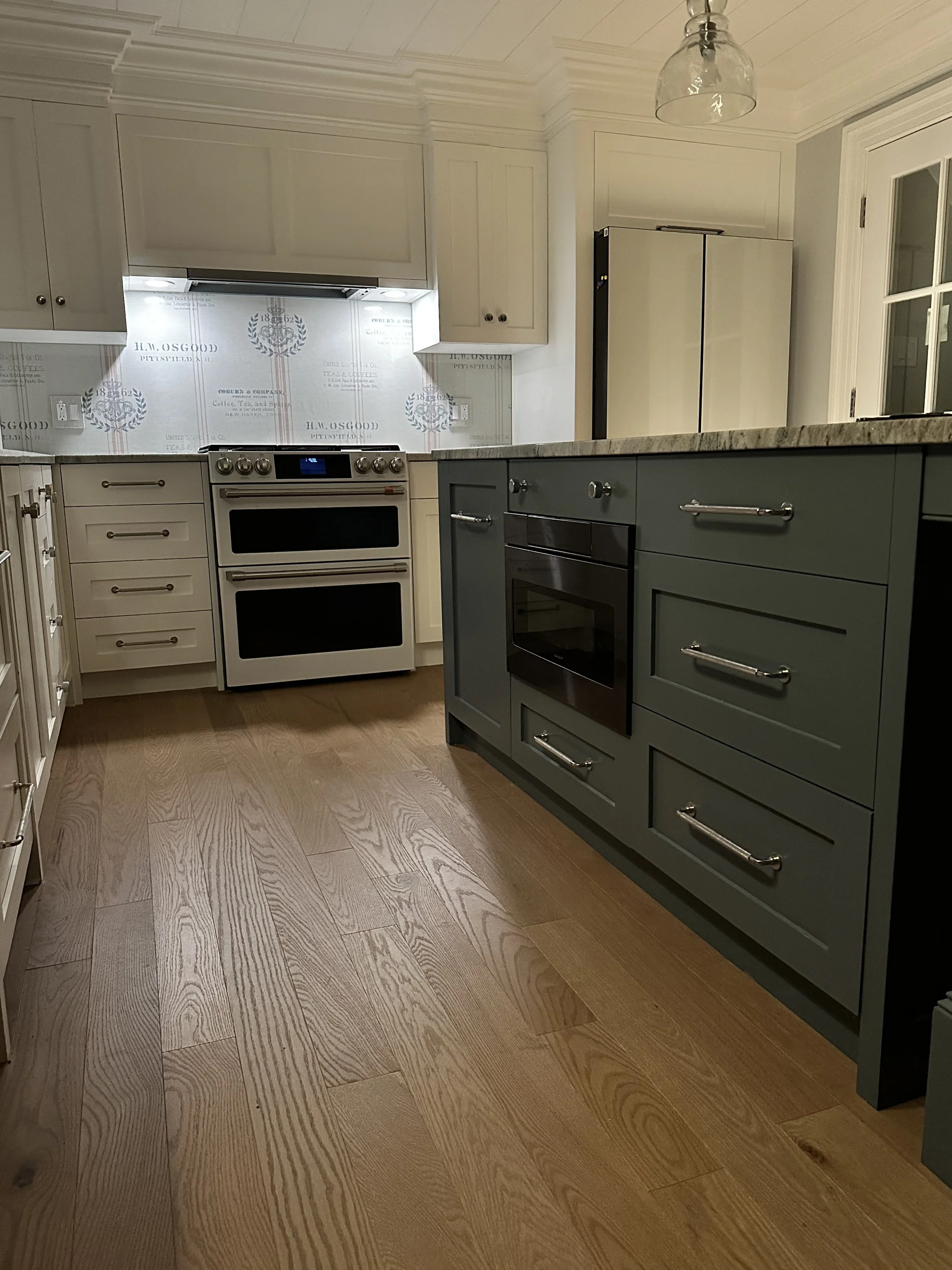A kitchen with white upper cabinets, a white stove, a gray island with drawers, and a black refrigerator. The floor is light wood and the ceiling is white with crown molding. The backsplash appears to be printed wallpaper with text and logos.