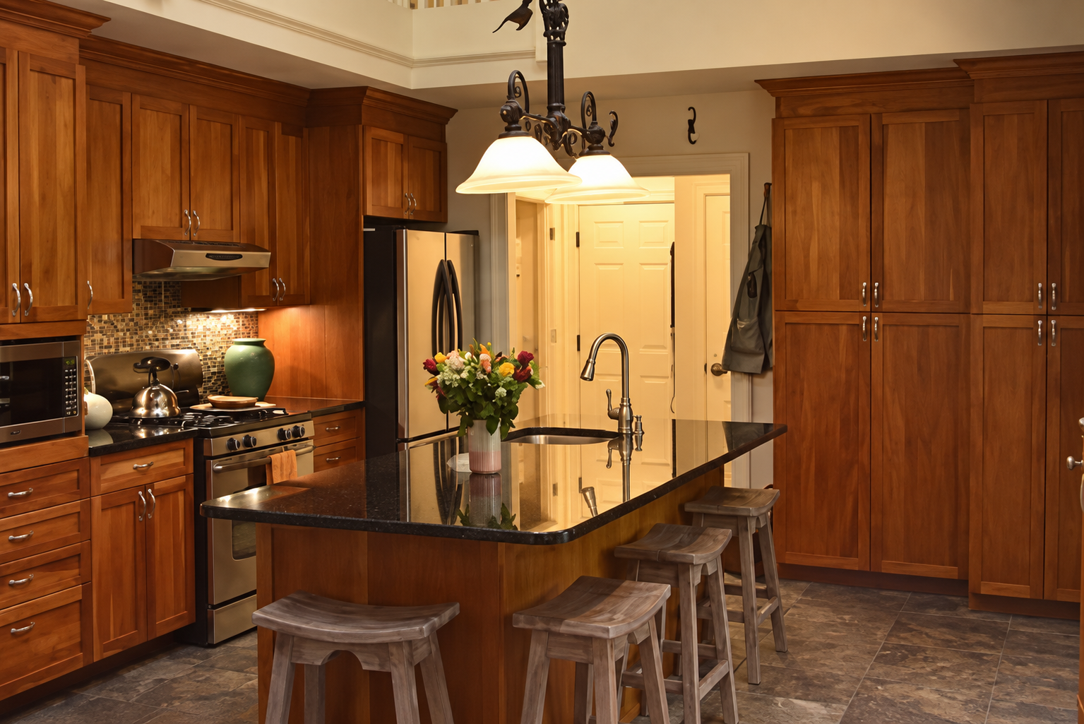 A kitchen with wooden cabinets, a black granite countertop, and a central island with a sink. There are four wooden bar stools at the island, a bouquet of flowers on the counter, and modern appliances including a stainless steel refrigerator and stov
