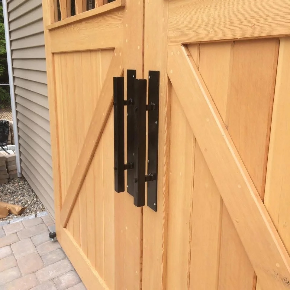 Close-up of a wooden outdoor gate with black hinges and hardware, surrounded by a wooden fence and brick pavement.