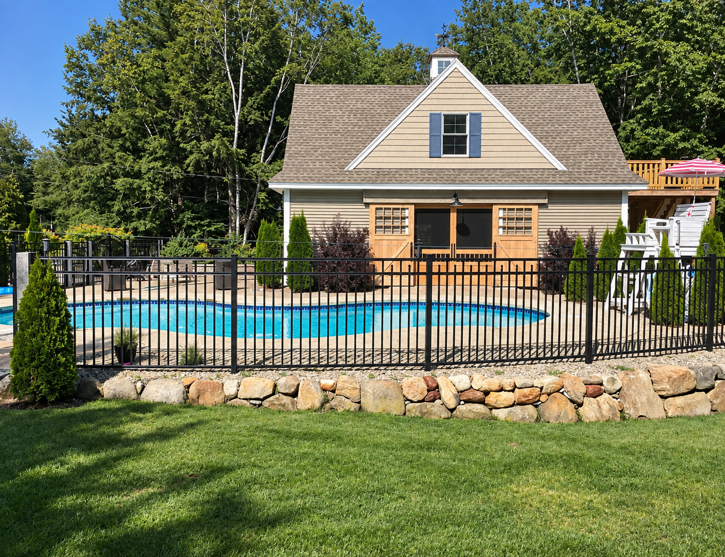 Backyard with in-ground swimming pool, surrounded by a black metal fence, a stone border, and a grassy lawn. A two-story house with beige siding, gable roof, and outdoor seating area with a deck and umbrella in the background.