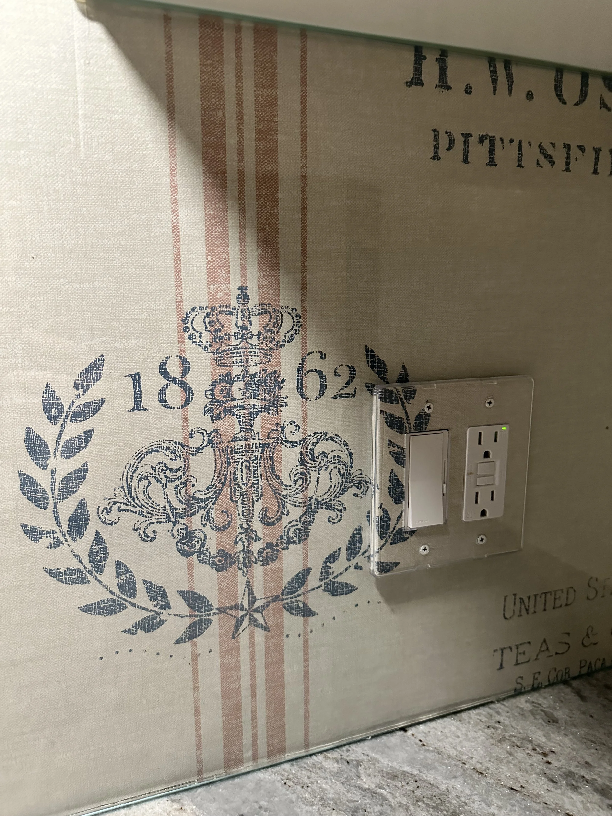 Blank wall with decorative wallpaper, electrical outlet, and light switch. The wallpaper features a vintage design with a crown and ornate patterns in shades of blue and beige.