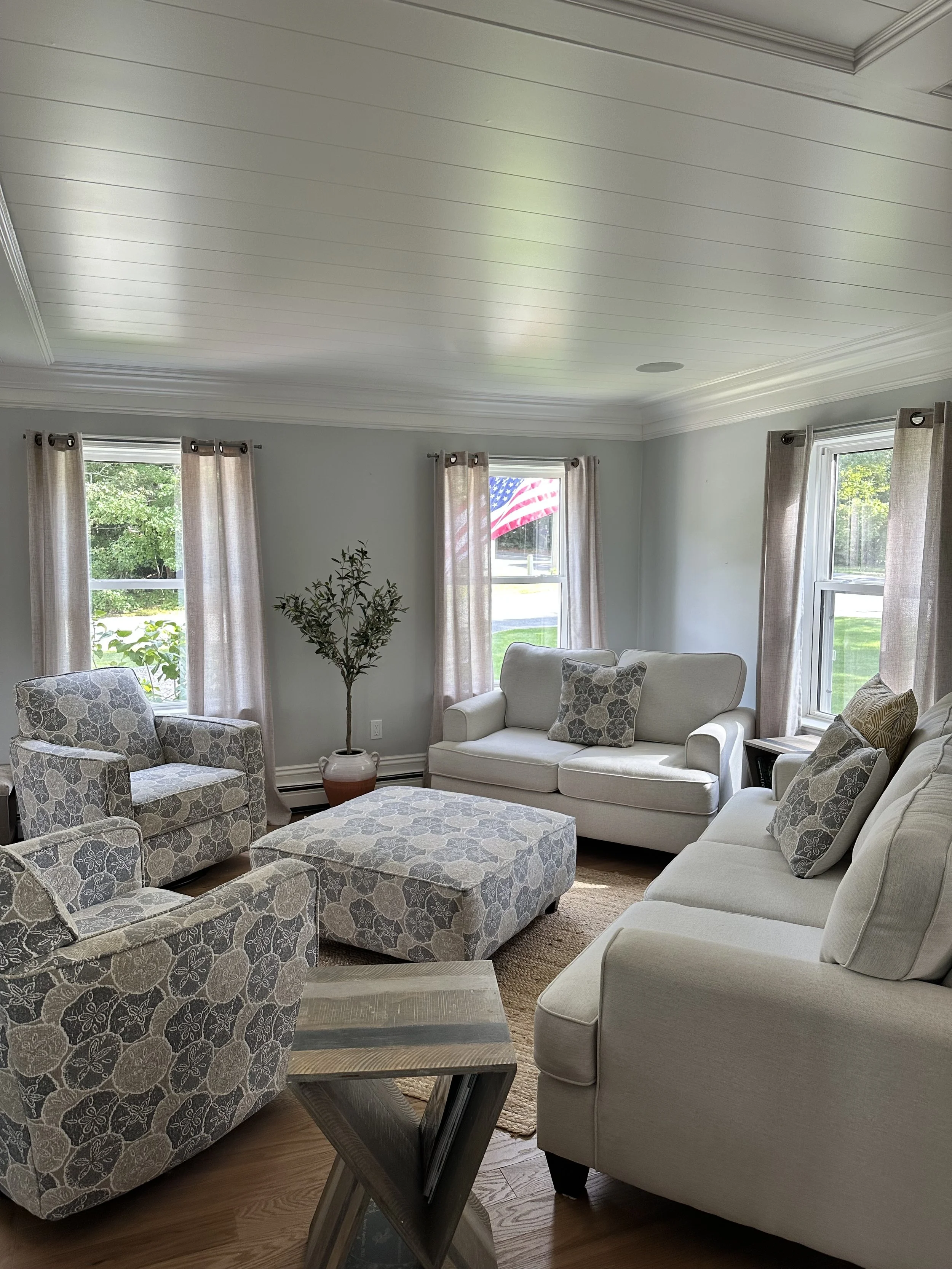 A cozy living room with three large windows, beige curtains, a white sofa with patterned pillows, an armchair with a similar pattern, an ottoman matching the armchair, a potted plant, a small side table, and an American flag visible through one window.