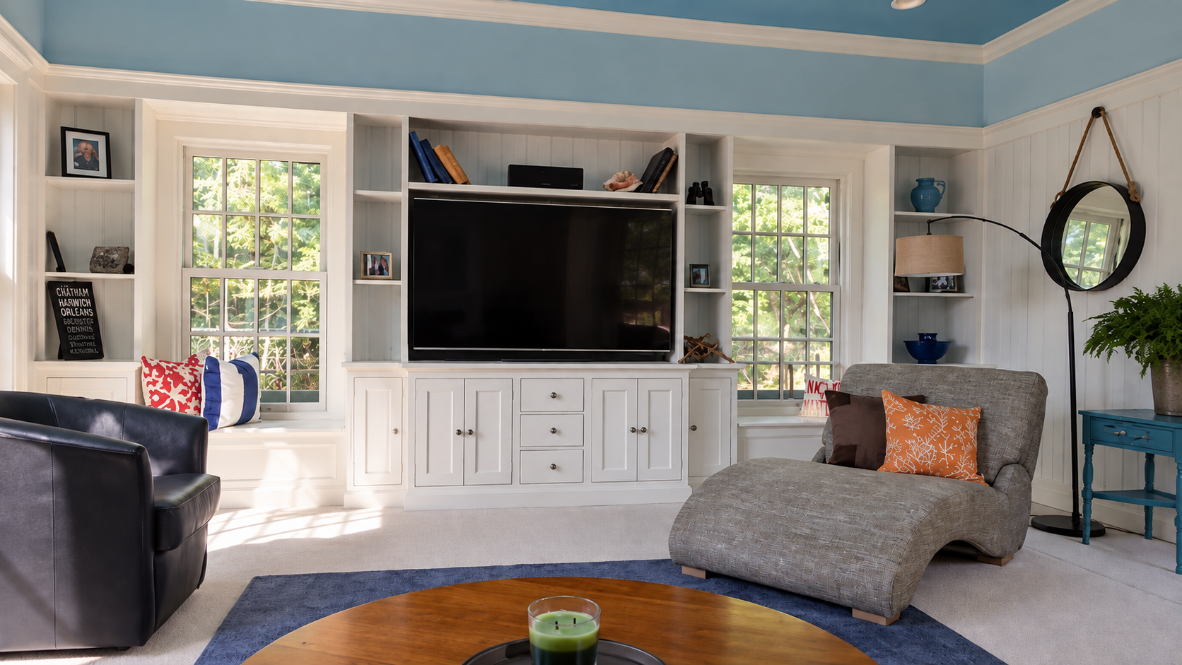 Living room with blue ceiling, white walls, and white built-in shelves. There are three windows with green outside views. A large flat-screen TV is centered in the shelving unit, with books, photographs, and decorative items. On the left, there is a 