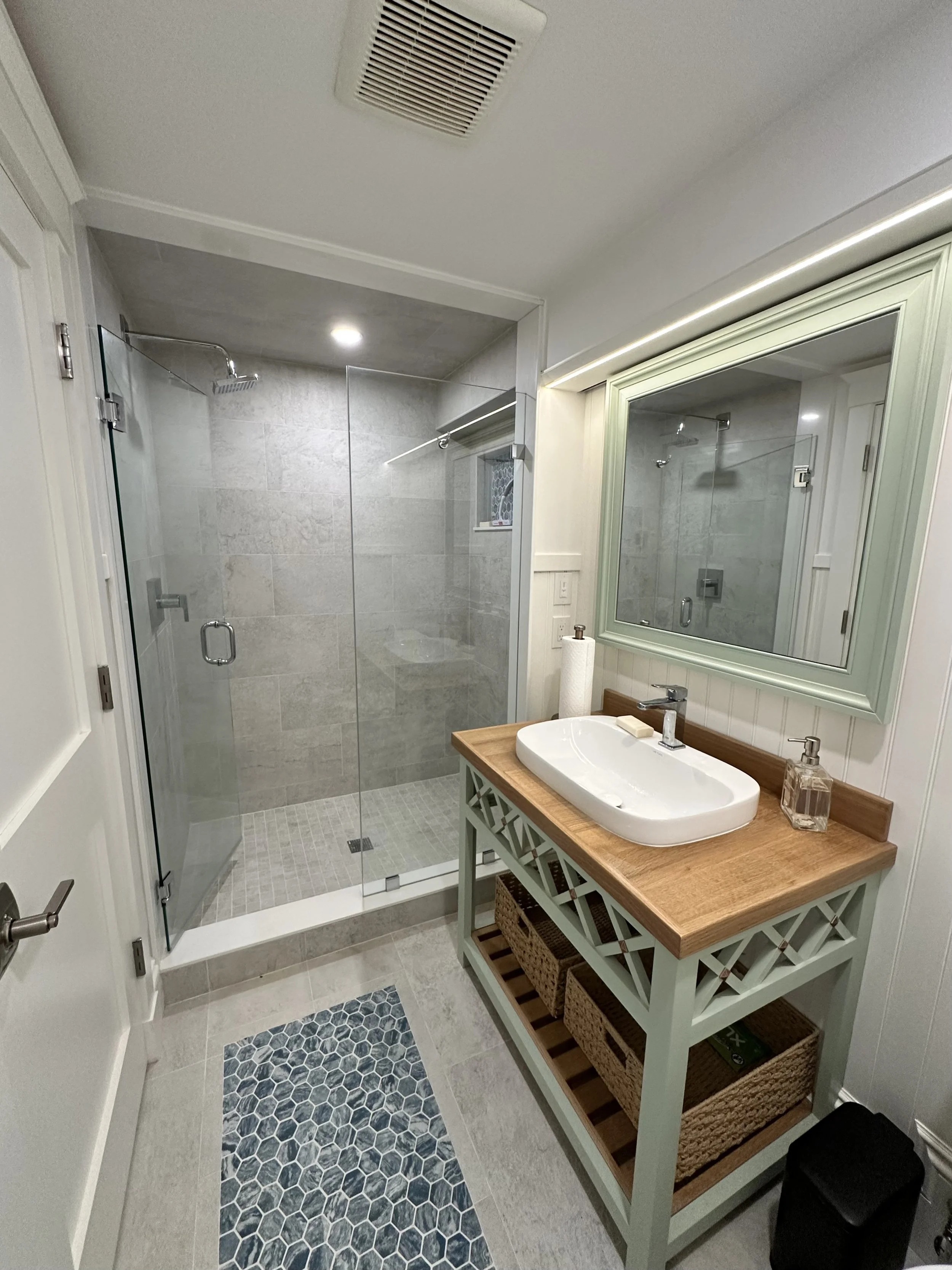 Bathroom with a walk-in shower enclosed by a glass door, a mirror framed in light green, a white vessel sink on a wooden countertop, and a cabinet with woven baskets underneath. The floor features hexagonal tiles, and there is a small window in the s