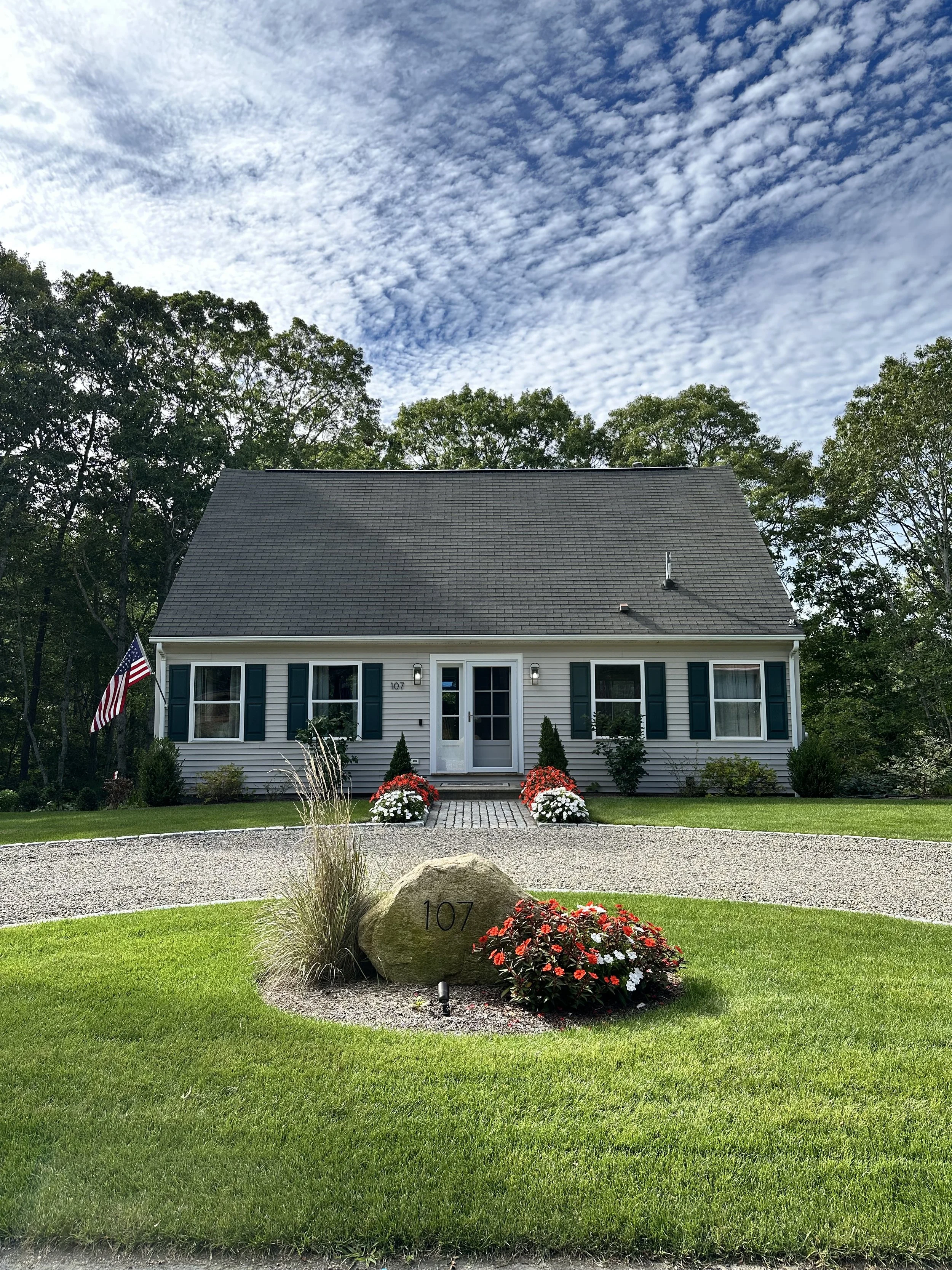 Front view of a house with a gray roof, white siding, green shutters, and a white front door. The yard features a rock with the house number 107 engraved, surrounded by red and white flowers and green grass. An American flag is displayed on a flagpol