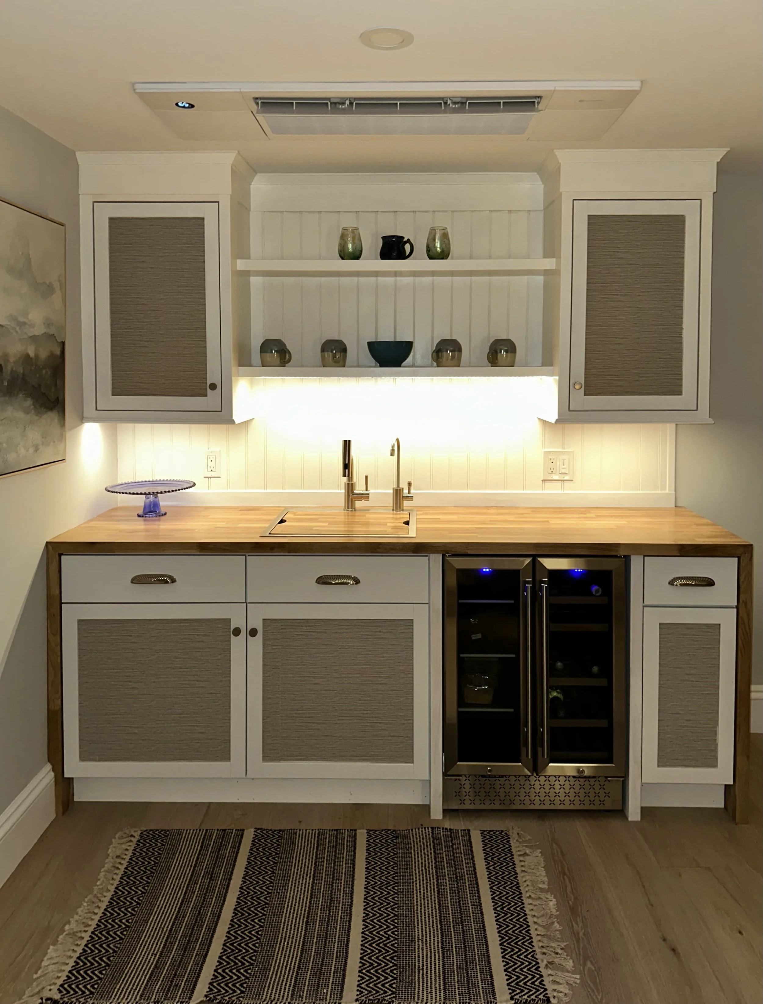 Kitchen with white cabinets, a wooden countertop, a built-in sink, a wine cooler, decorative vases on open shelving, a purple decorative tray, and a patterned rug on hardwood floors.