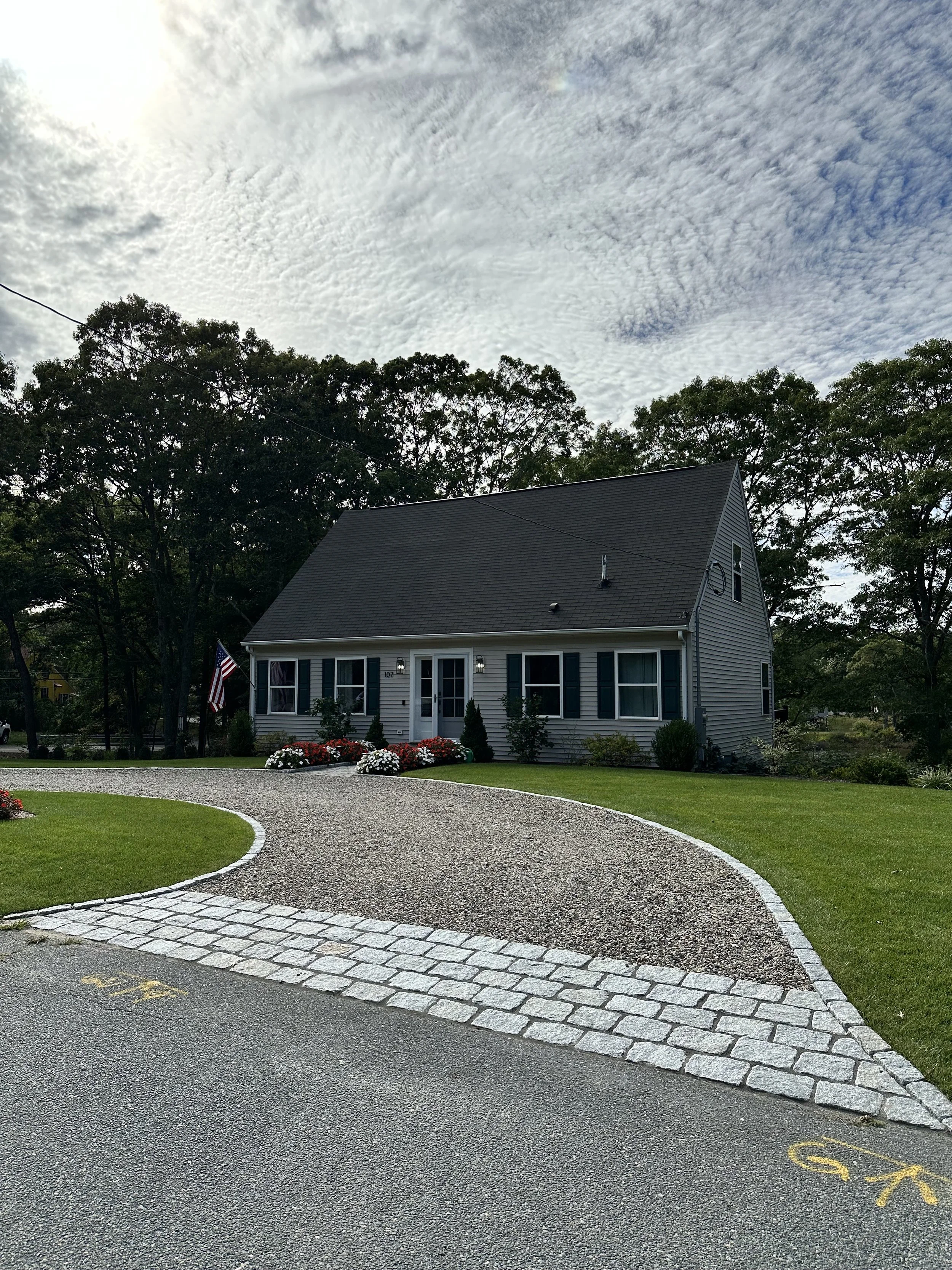Residential house with a gray roof, white siding, and black shutters, surrounded by a well-maintained lawn and flower beds. The driveway is curved and edged with cobblestones, and it leads to a street with yellow parking markings. The sky is partly c