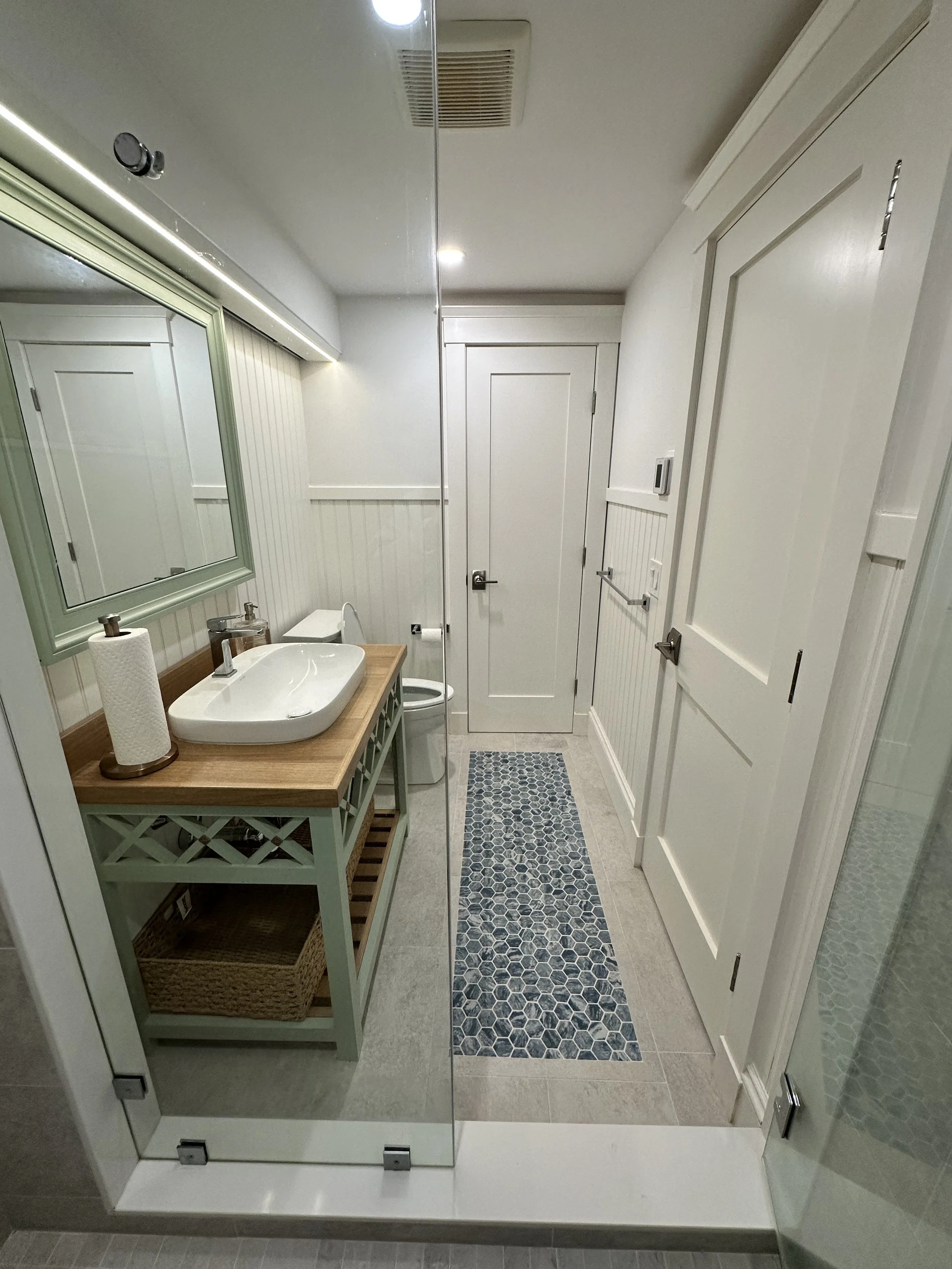A bathroom with a white door, a small sink with a wooden countertop, a mirror, a navy and white hexagon tile rug, beige tiled flooring, and various white built-in cabinets and walls.