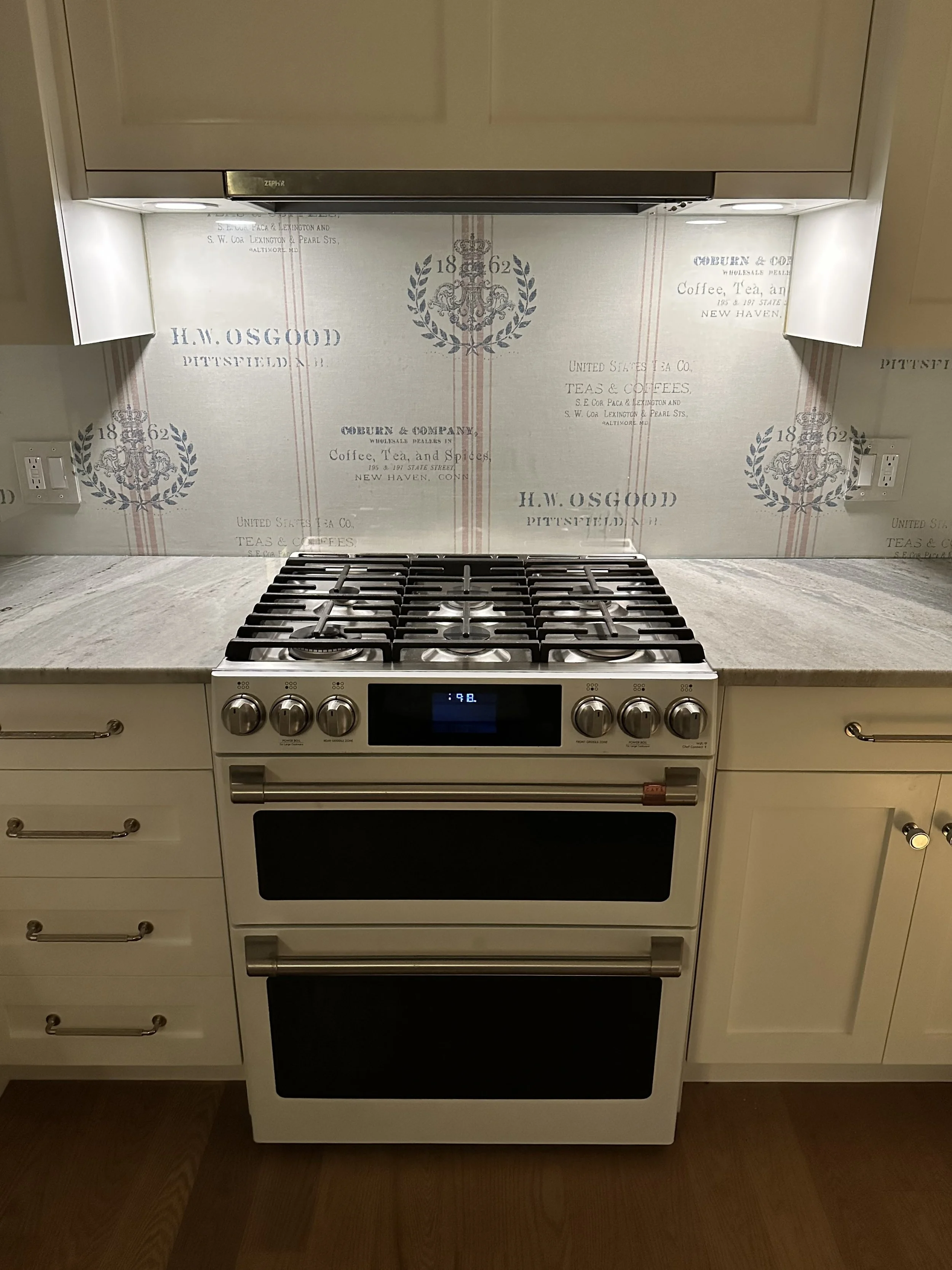 White kitchen stove with control knobs, set in white cabinets, with a vintage inspired wallpaper behind featuring the text 'H.W. Osgoode, Pittsfield, MA, 1862' and other vintage-style labels.