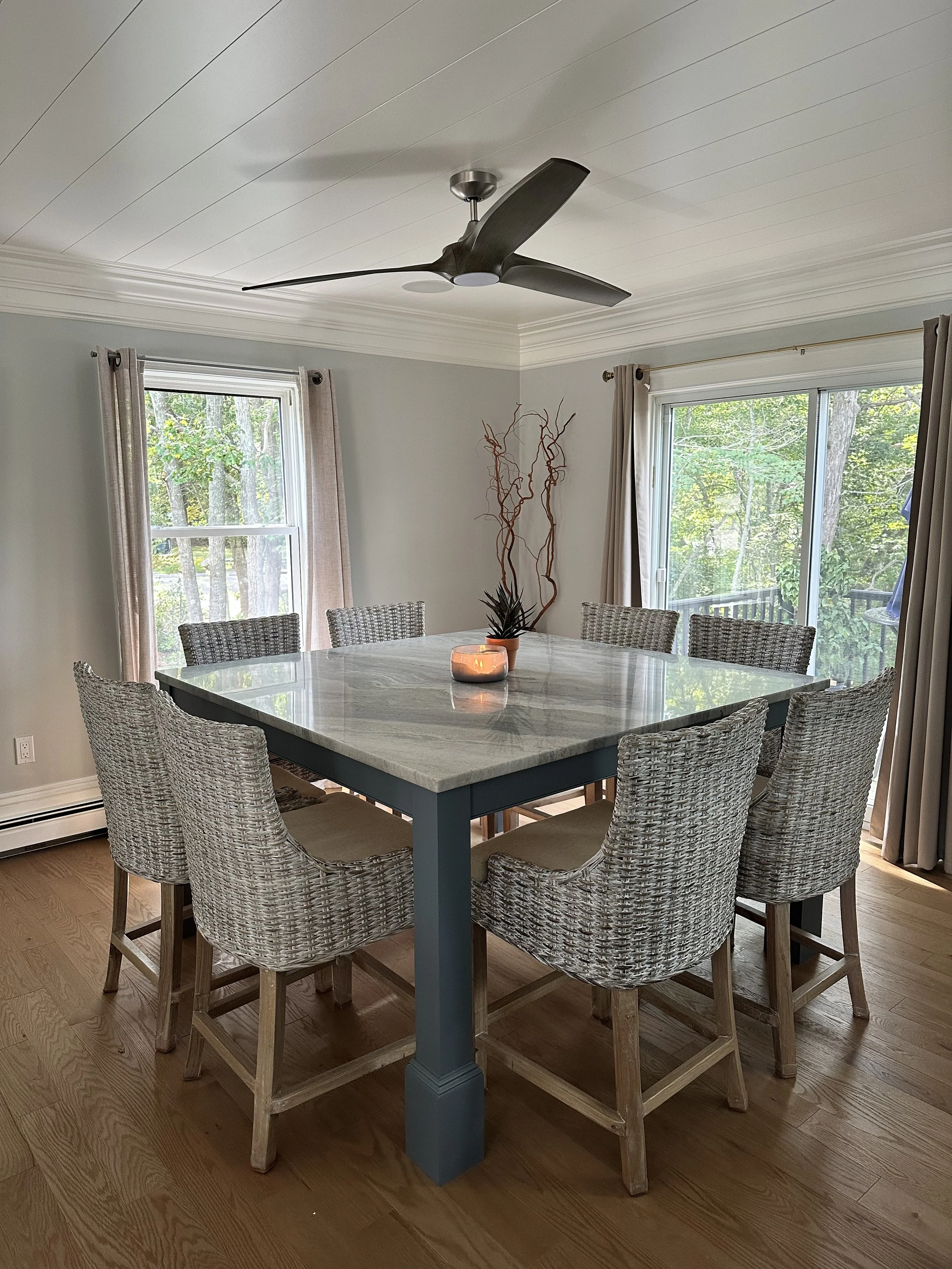 A dining room with a marble table surrounded by wicker chairs, large windows with curtains, a ceiling fan, and a decorative vase with branches on the table.