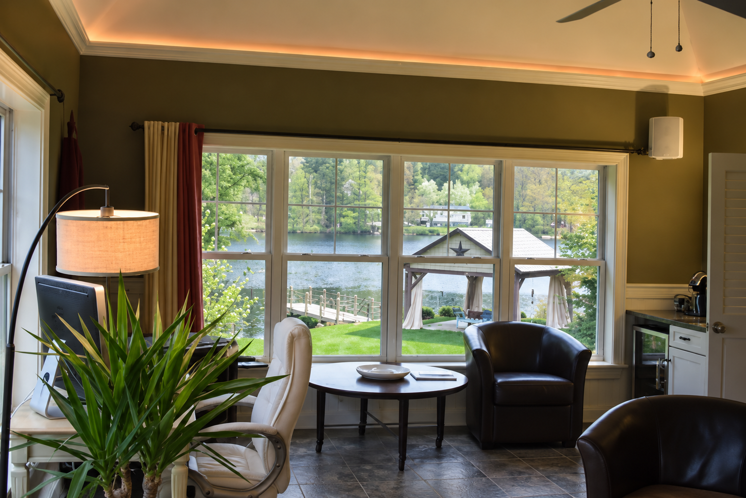 Living room with large window overlooking a lake, with a small wooden dock and gazebo outside. Inside, there's a desk with a computer, a white chair, a floor lamp, a round table, and leather armchairs.