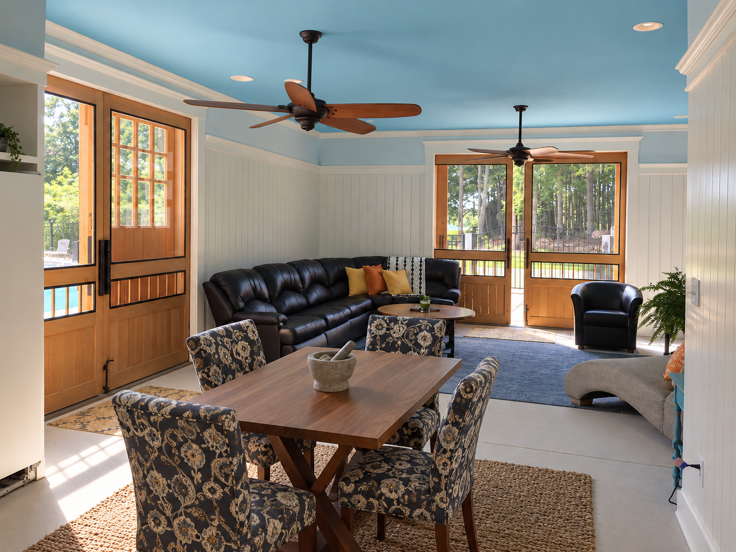 Living room with blue ceiling, white paneled walls, wooden doors and large windows, black leather sofa and armchair, round coffee table with plant, patterned dining chairs and table, ceiling fans, and outdoor view.