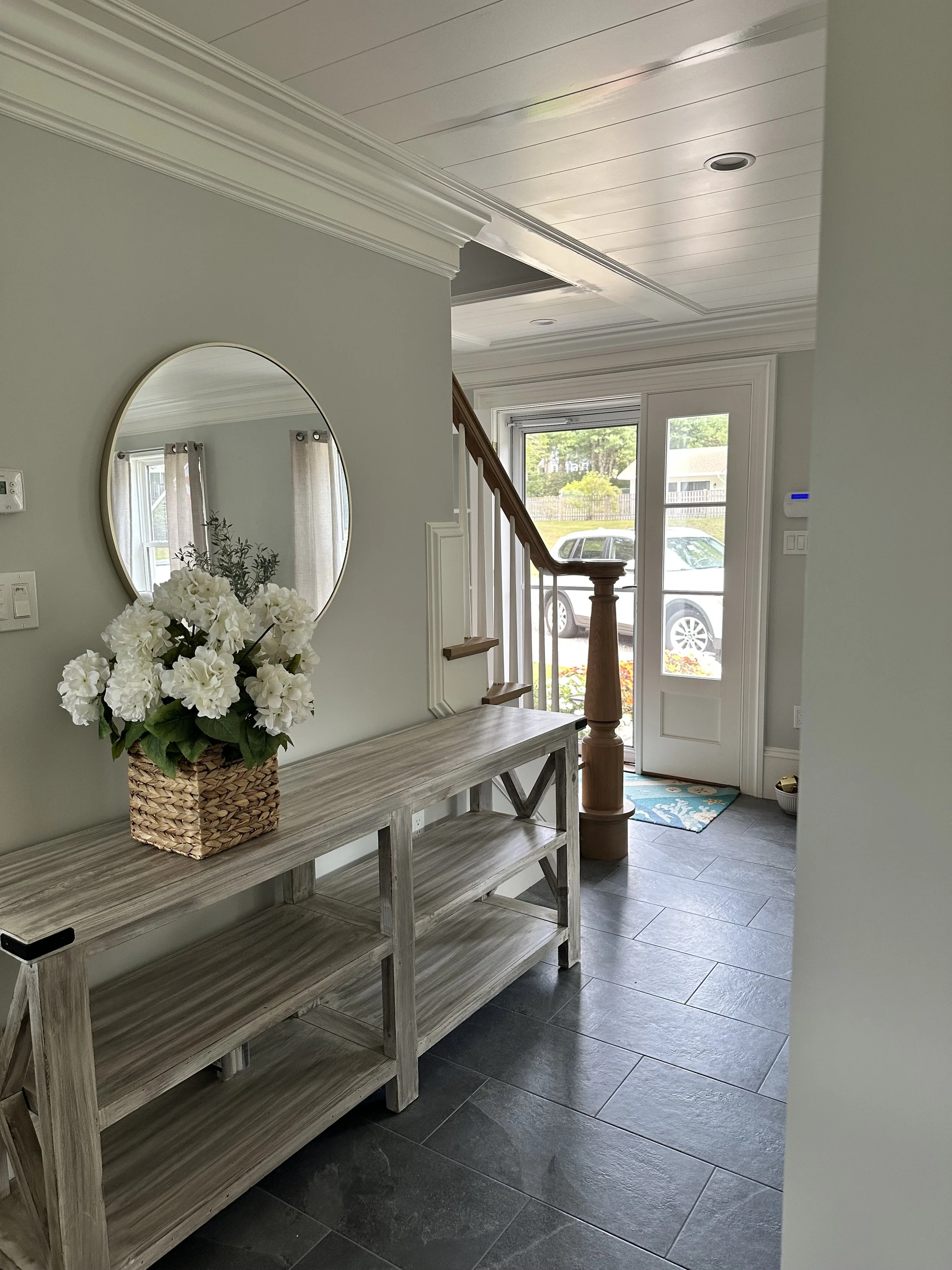 Entryway with a wooden console table, a round mirror on a light gray wall, a vase of white flowers, a staircase with wooden railing, and a front door with windows showing a car and greenery outside.