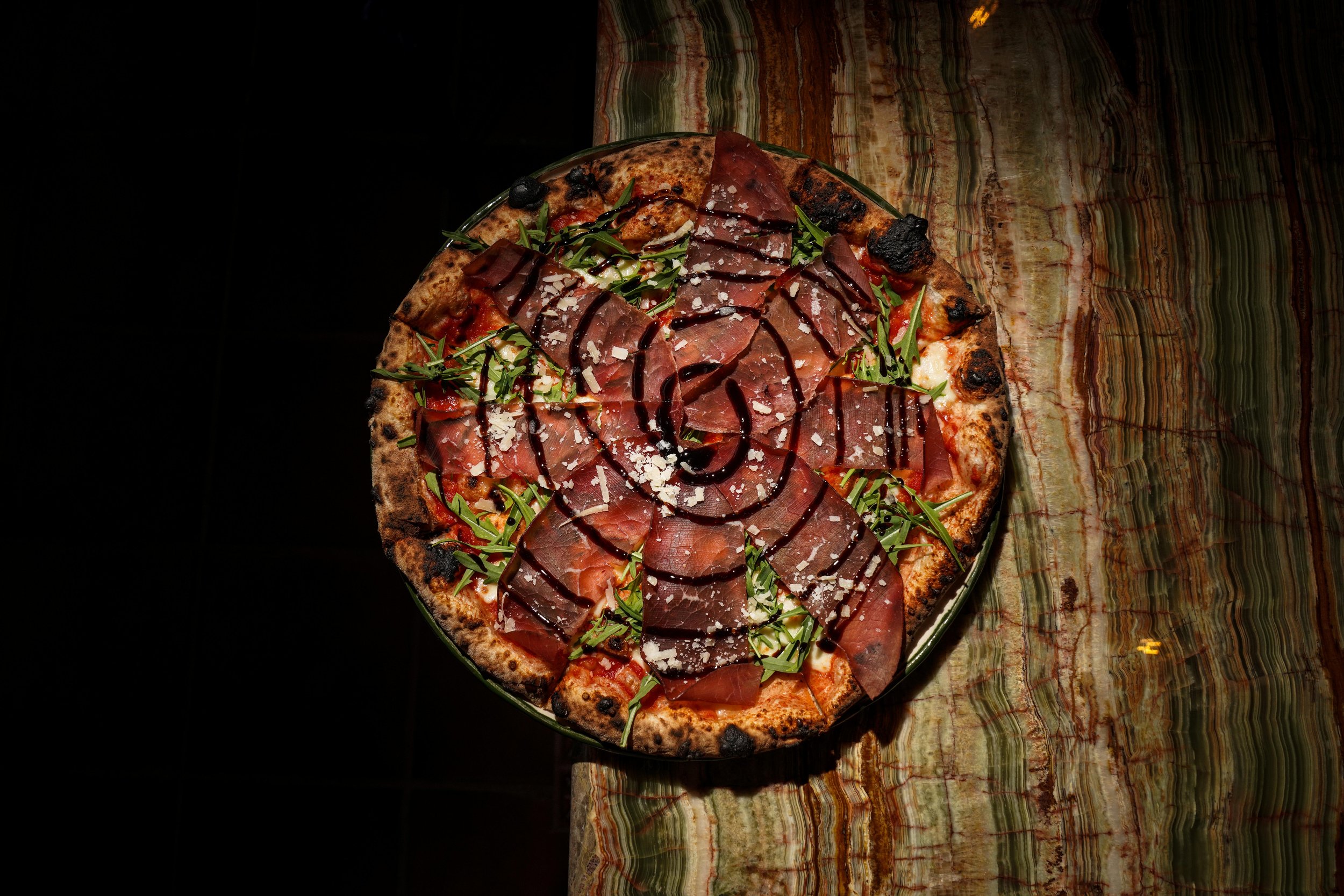 A pizza topped with slices of cured meat, arugula, grated cheese, and a drizzle of balsamic glaze, served on a colorful marble table.