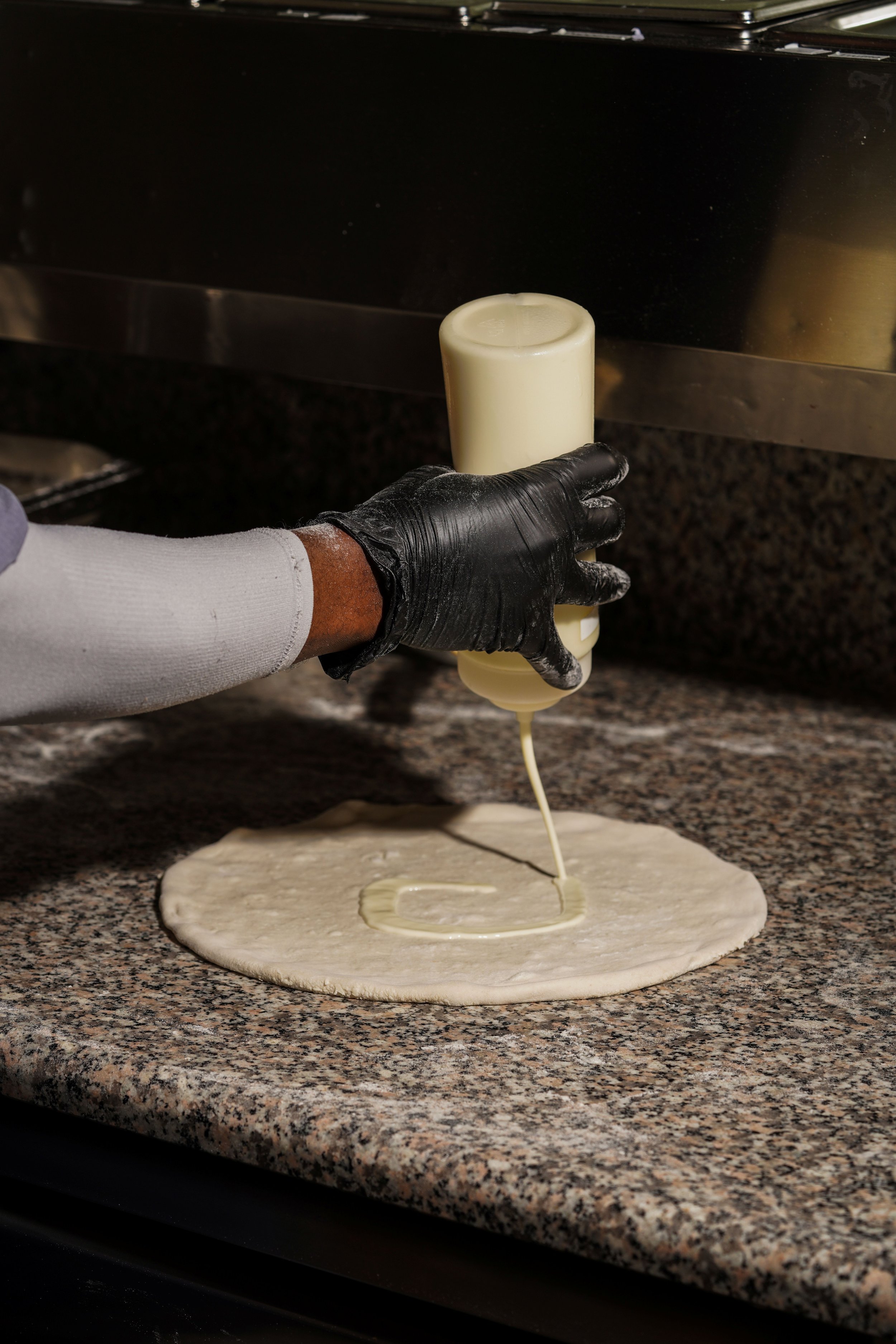 A person in a black glove and a long sleeve shirt is holding a squeeze bottle of off-white sauce over pizza dough on a granite countertop, preparing to make a pizza.
