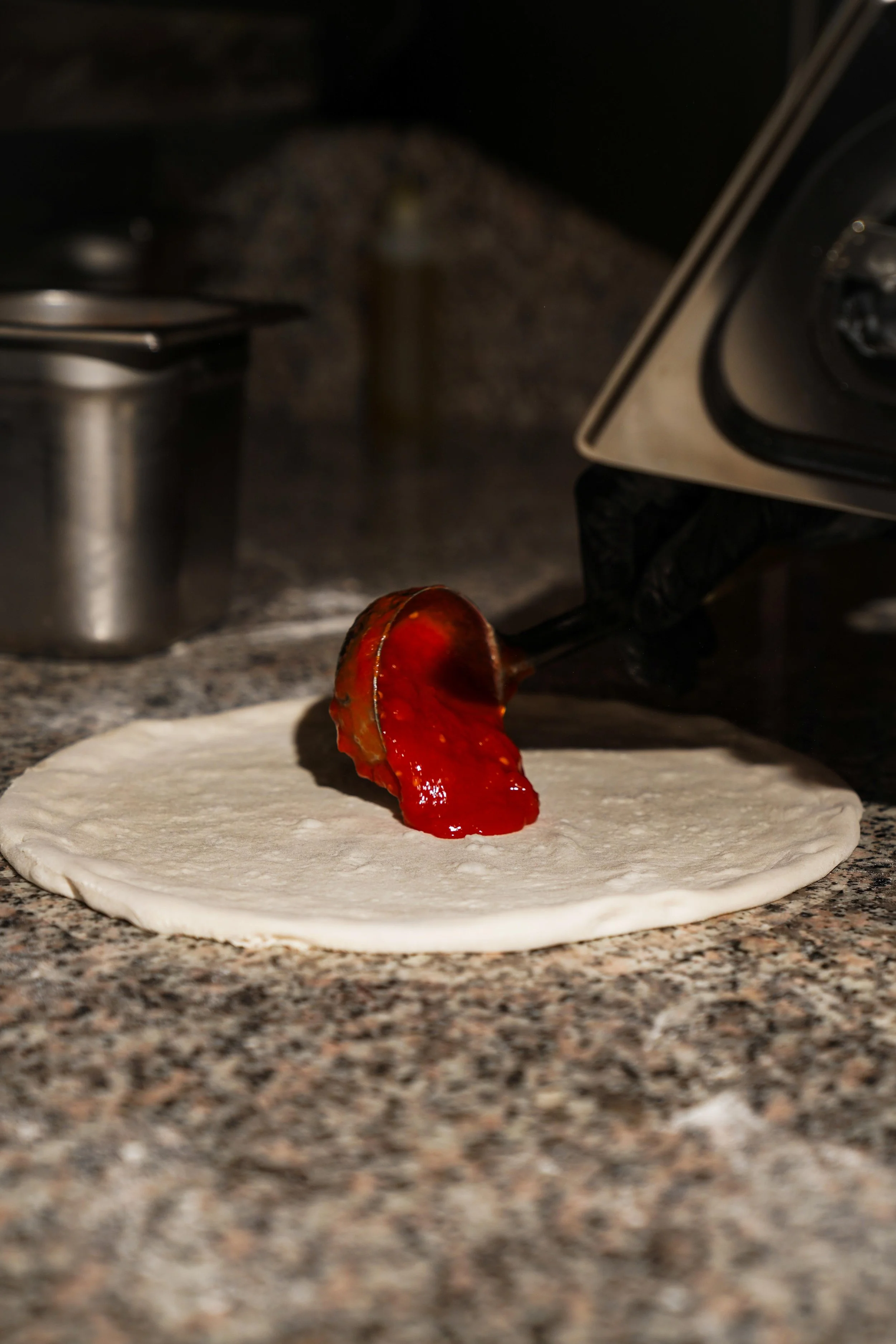 A person adding red sauce to a rolled out pizza dough on a granite countertop, with a stove and coffee pot in the background.