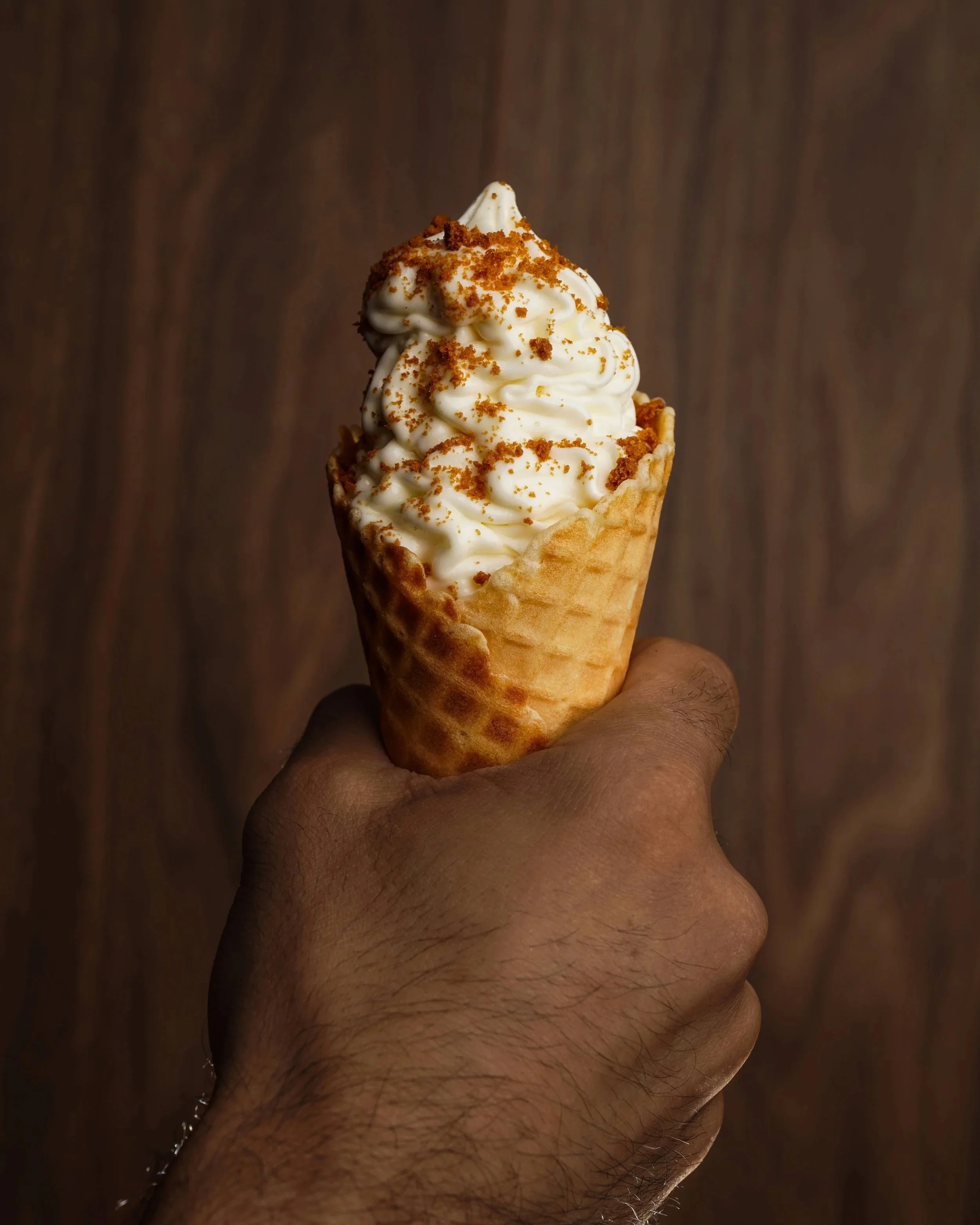 A hand holding a soft-serve vanilla ice cream cone with a waffle cone, topped with crushed cookies or graham crackers, against a wooden background.