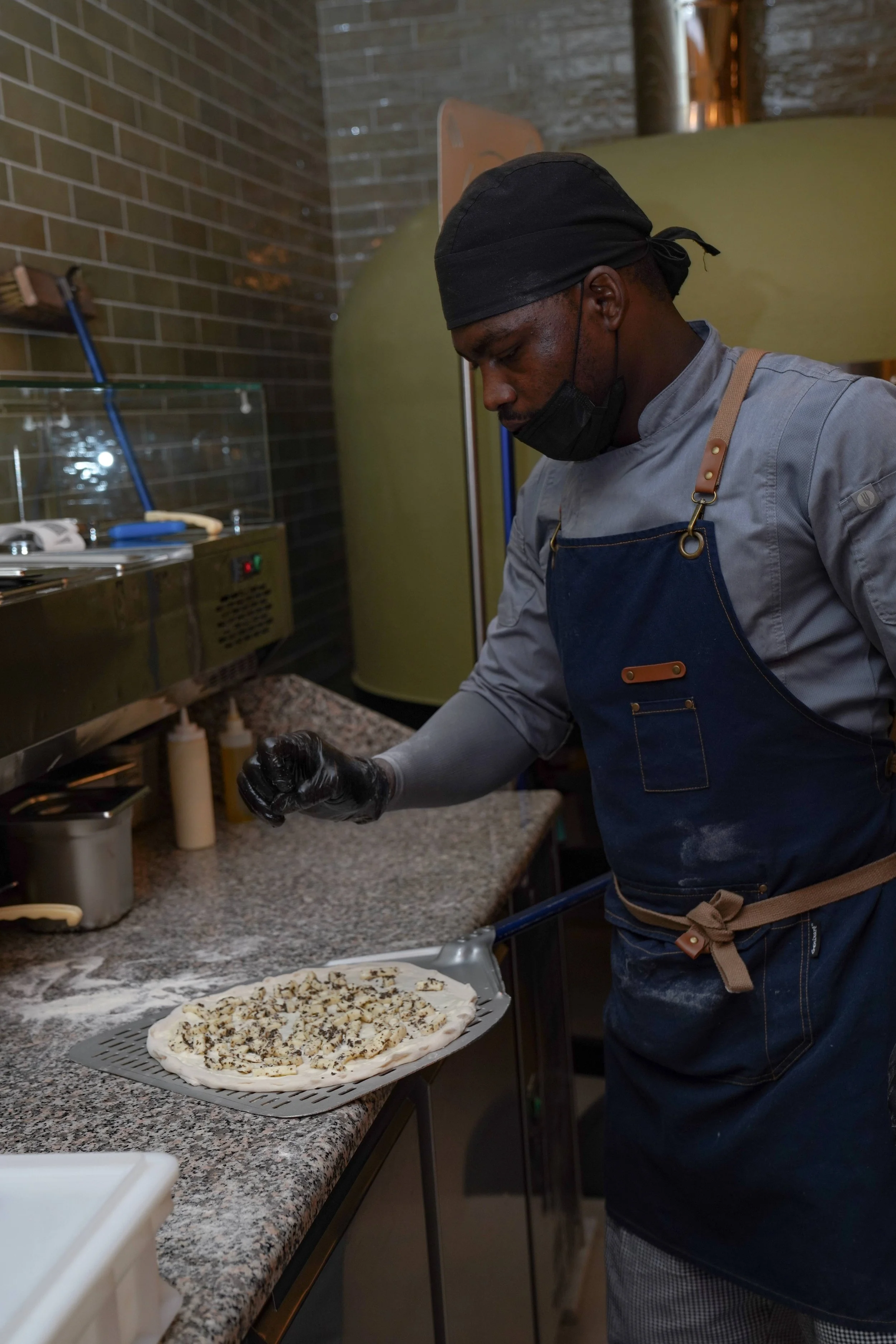 A person wearing a black headscarf, black face mask, gray chef coat, and navy apron is preparing a pizza with diced toppings at a kitchen counter. The pizza is on a tray and garnished with various ingredients.